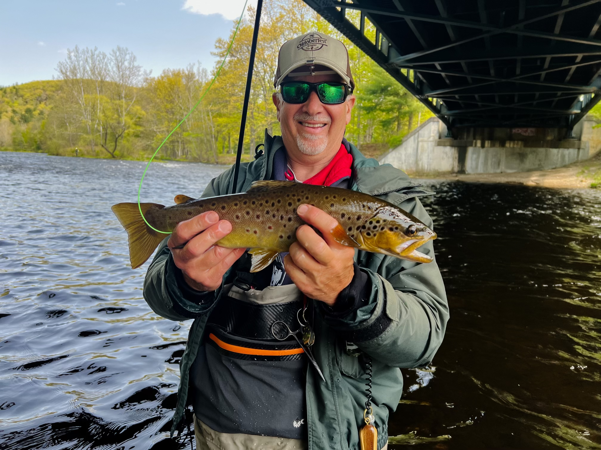 middle-aged man smiling, wearing tan hat, and wearing sunglasses while standing in water and holding fish horizontally