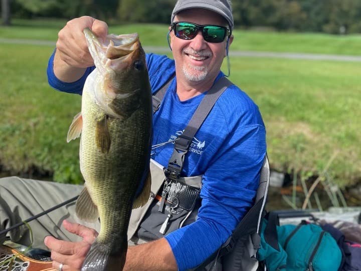 middle-aged man sitting on boat, wearing sunglasses, wearing a baseball hat, smiling, and holding a fish vertically