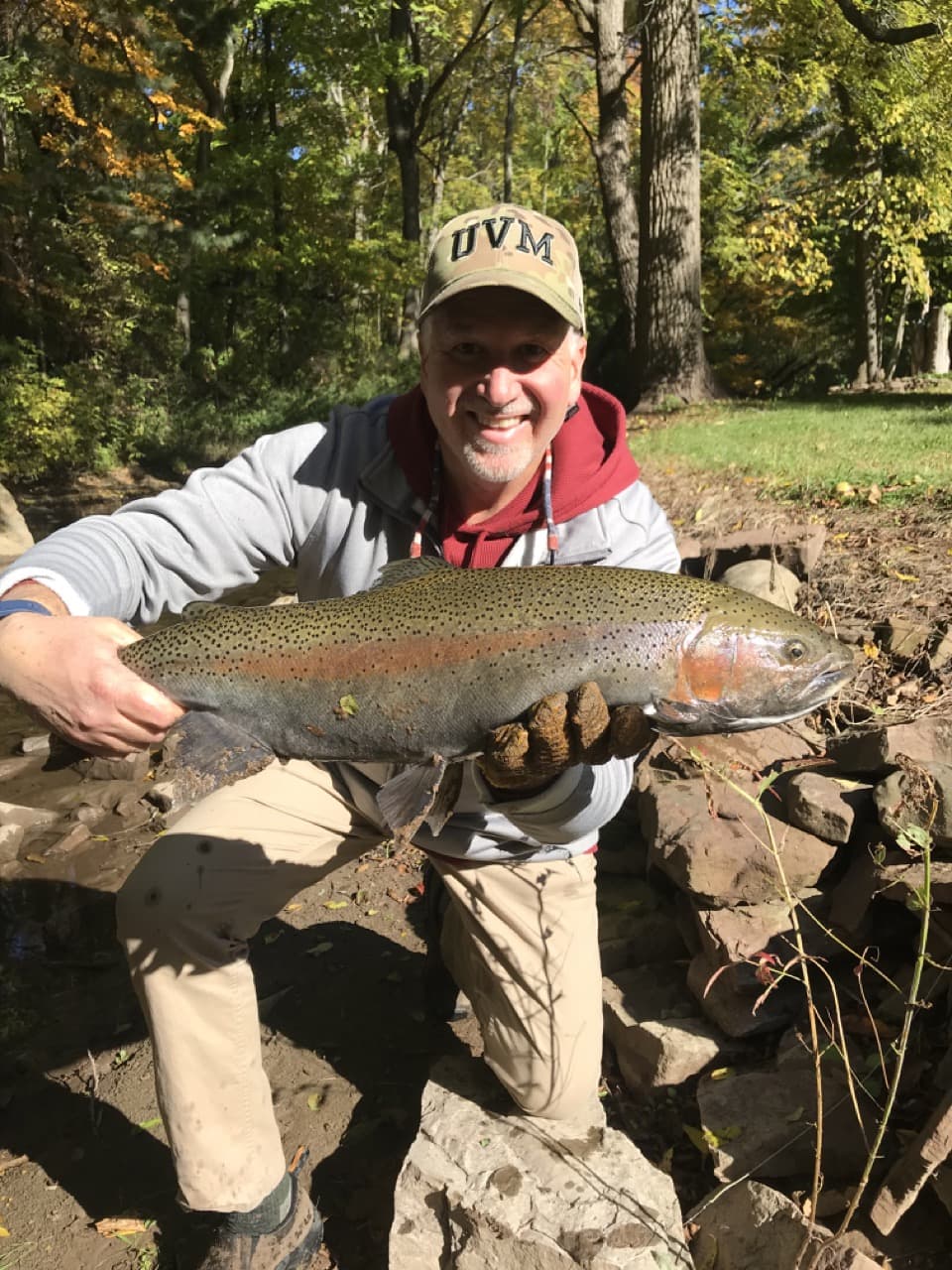 middle-aged man on one knee, wearing a grey jacket with red sweatshirt, wearing a baseball UVM hat, smiling, and holding a fish horizontally