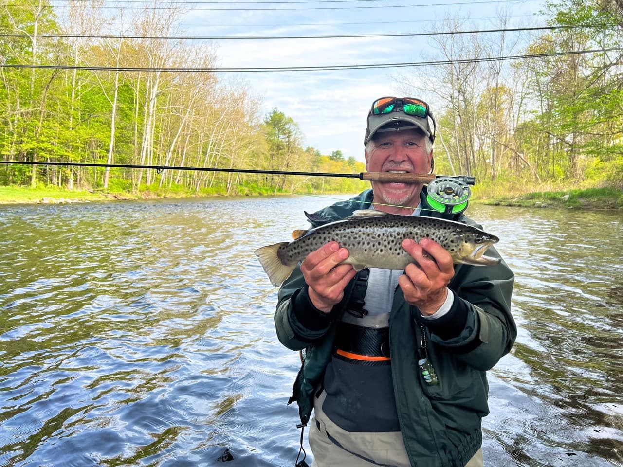 middle-aged man starnding in water, wearing a green jacket, wearing a baseball hat with sunglasses on top, biting a fishing rod handle, and holding a fish horizontally