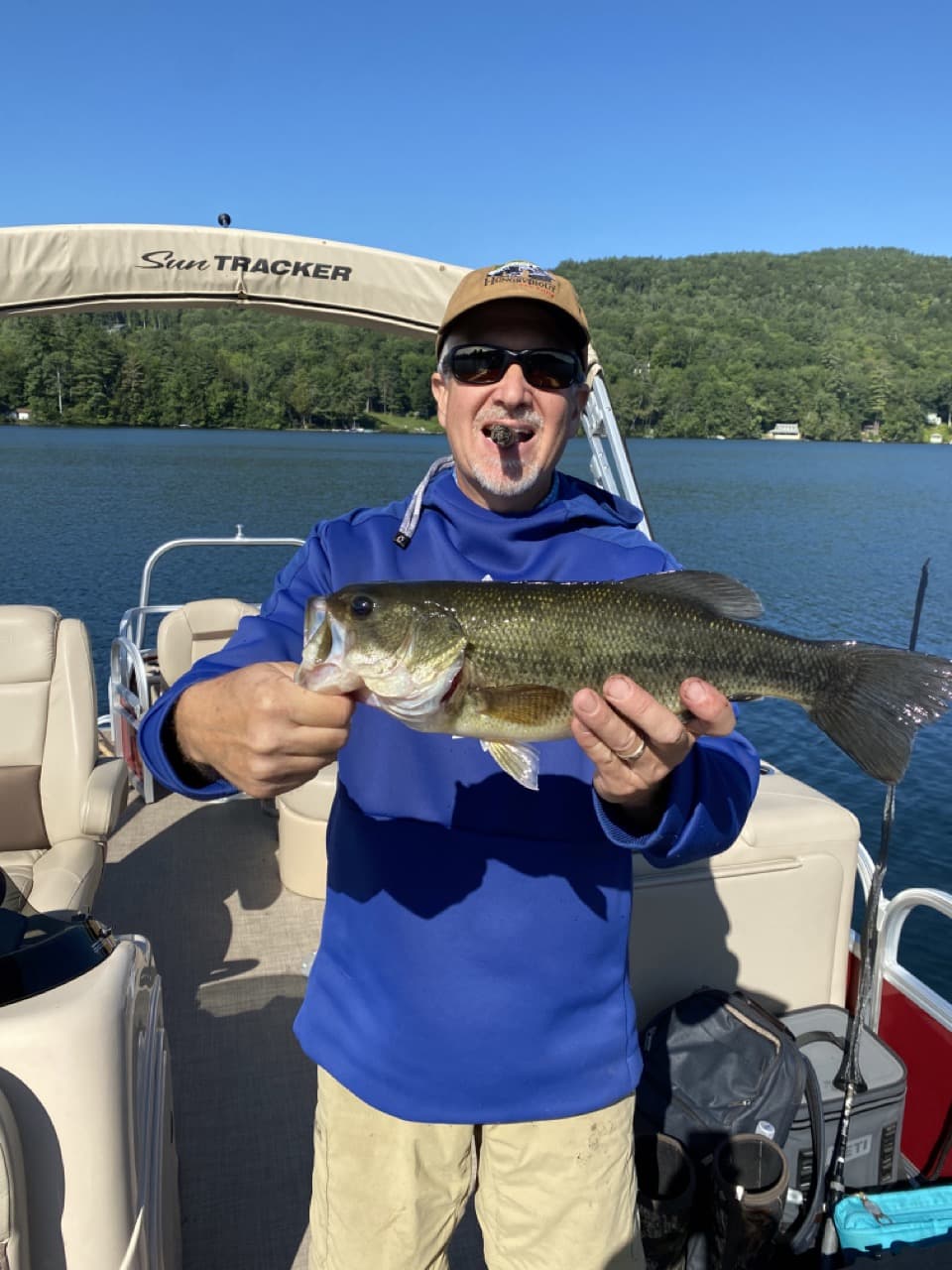 middle-aged man standing on a pontoon boat, wearing a blue sweatshirt, wearing a baseball hat, wearing sunglasses, biting a cigar, and holding a fish horizontally