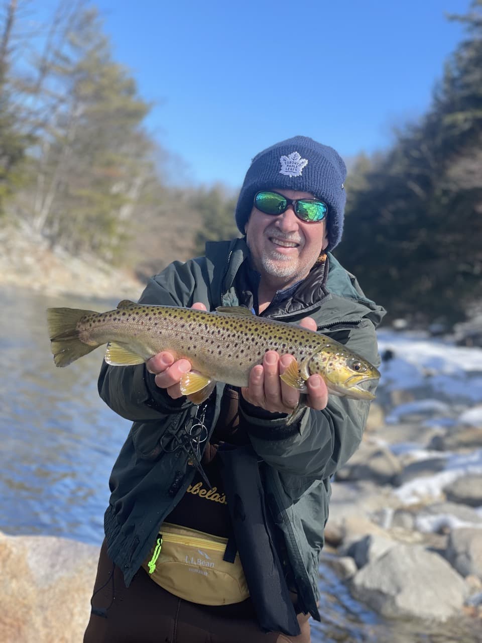 middle-aged man standing beside a river, wearing a winter green jacket, wearing a winter hat, wearing sunglasses, smiling, and holding a fish horizontally