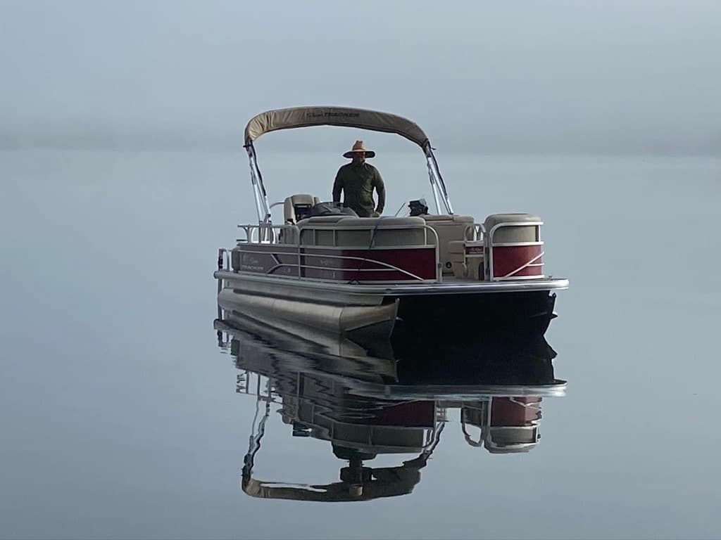 Man standing on a red and white pontoon boat in the middle of a foggy lake 