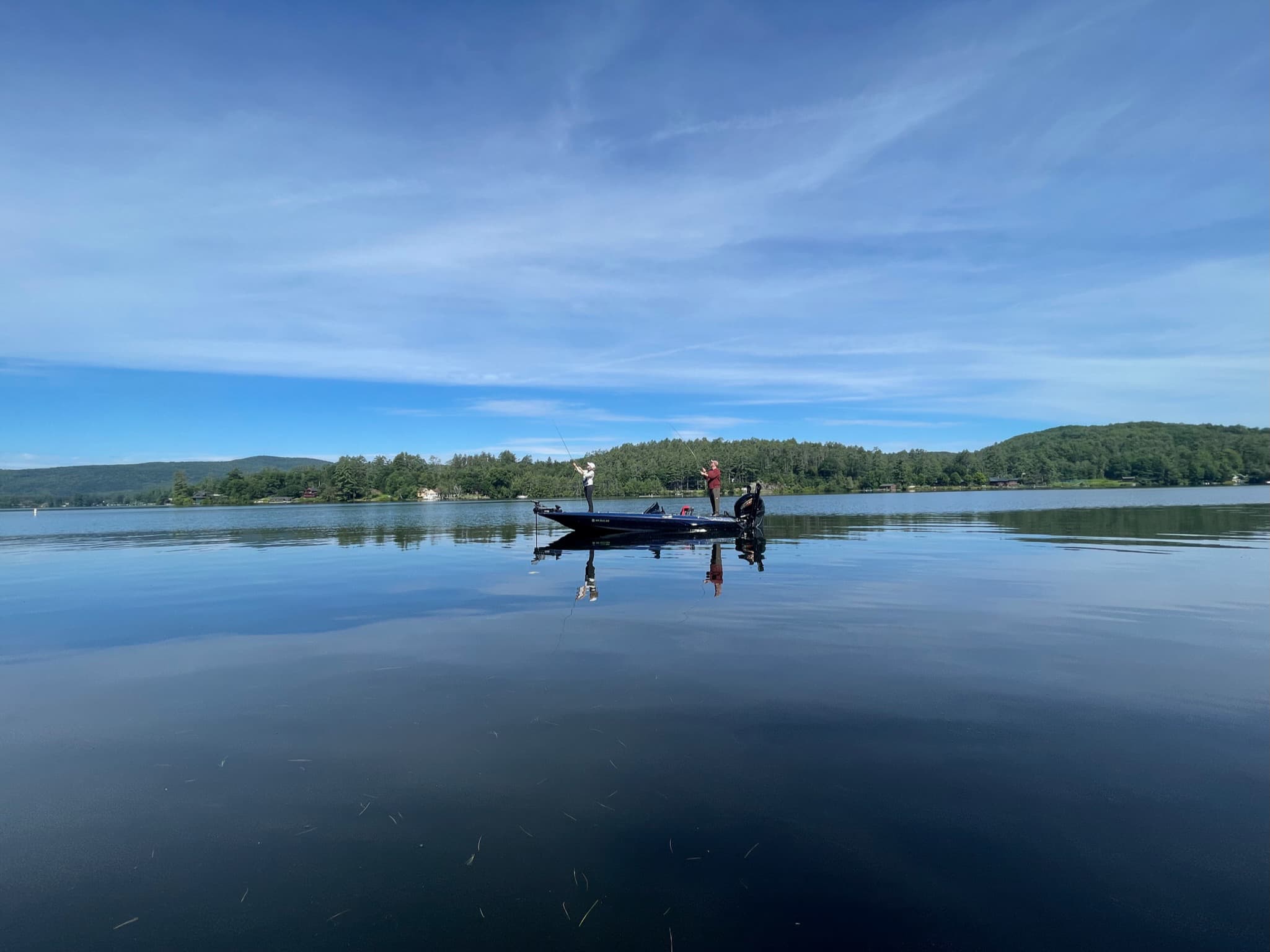 Zoomed out photo of man in red shirt and woman in white jacket and blank pants fly fishing on blue boat