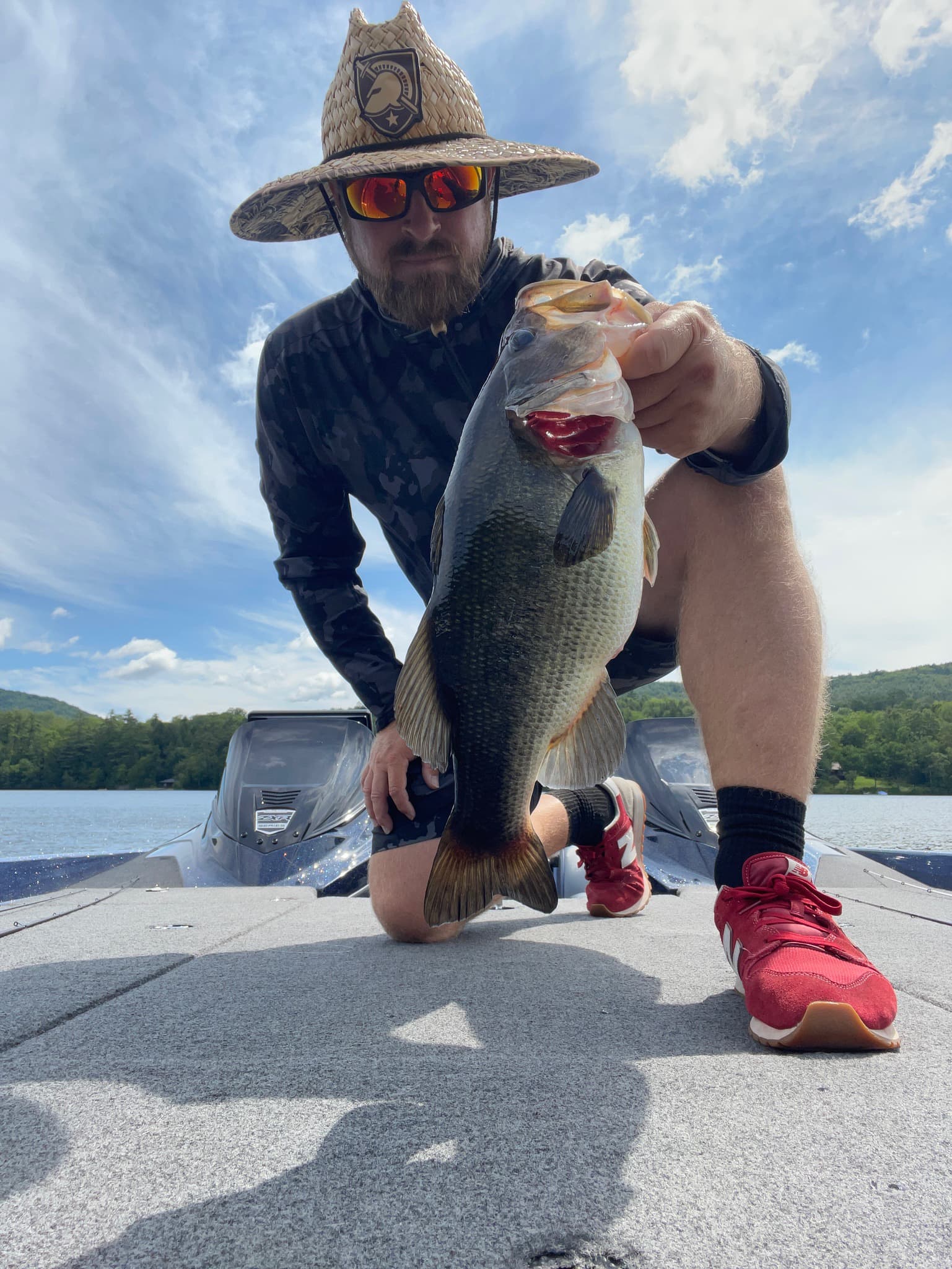 Man in straw hat and black shirt kneels down holding largemouth bass on boat