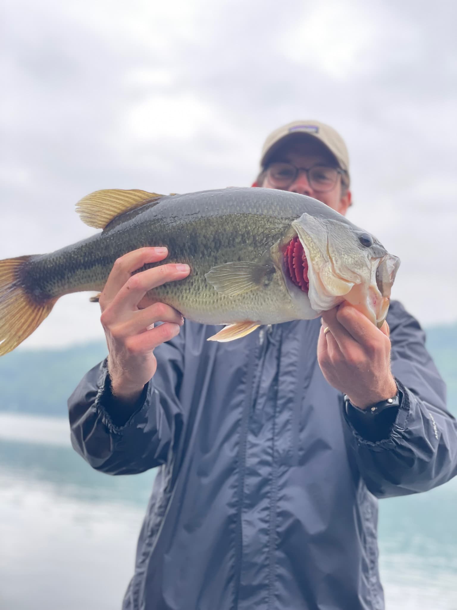 man wearing black jacket, glasses, and beige baseball cap holding up fish with gills out