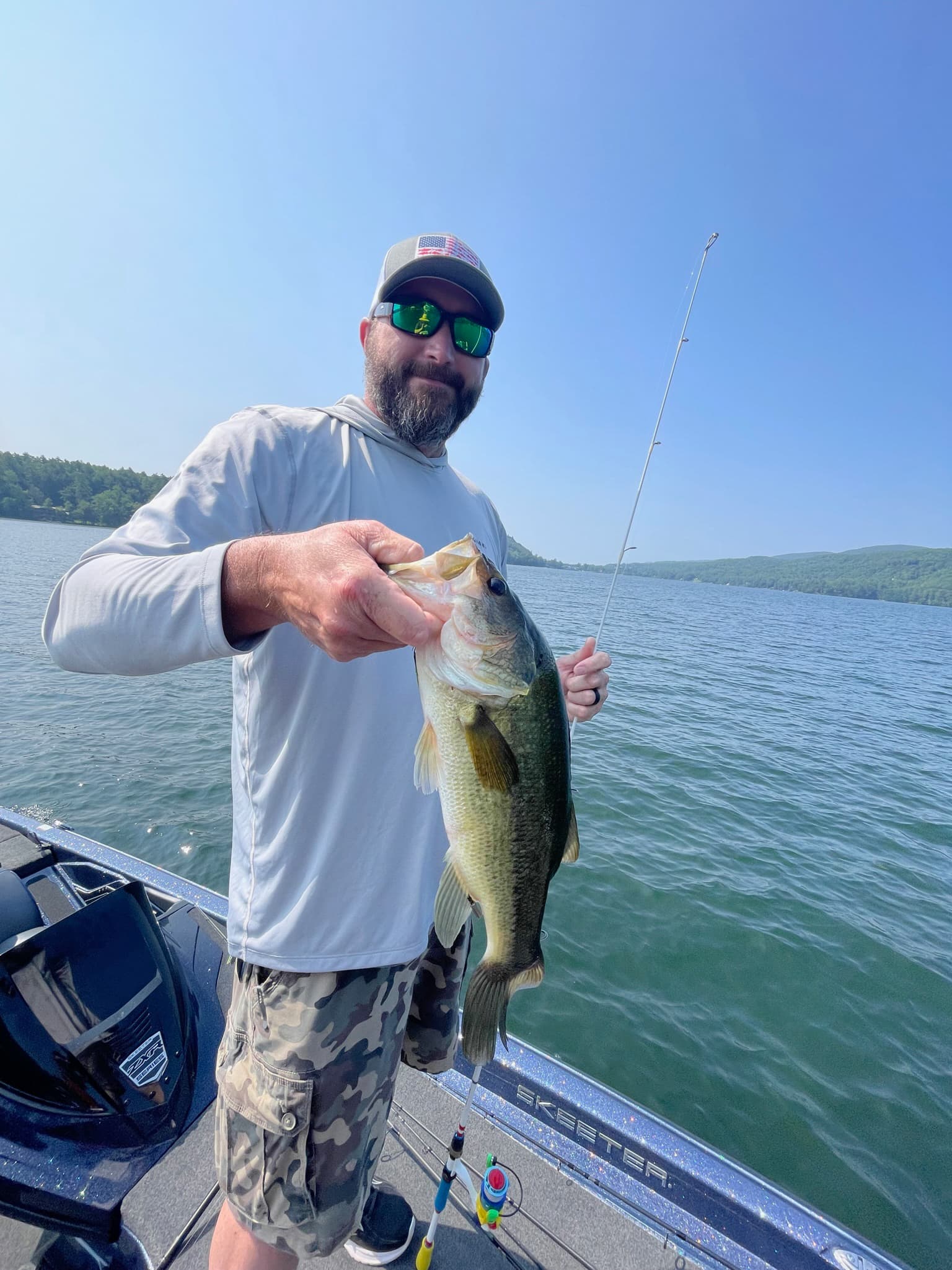 Man in green sunglasses, grey shirt, and camo shorts holds largemouth bass on blue boat