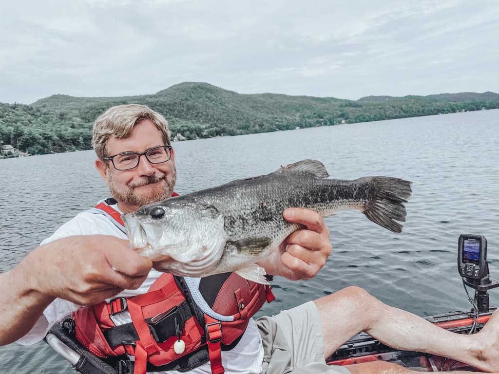 Man in white shirt and glasses holding one fish while smirking in kayak