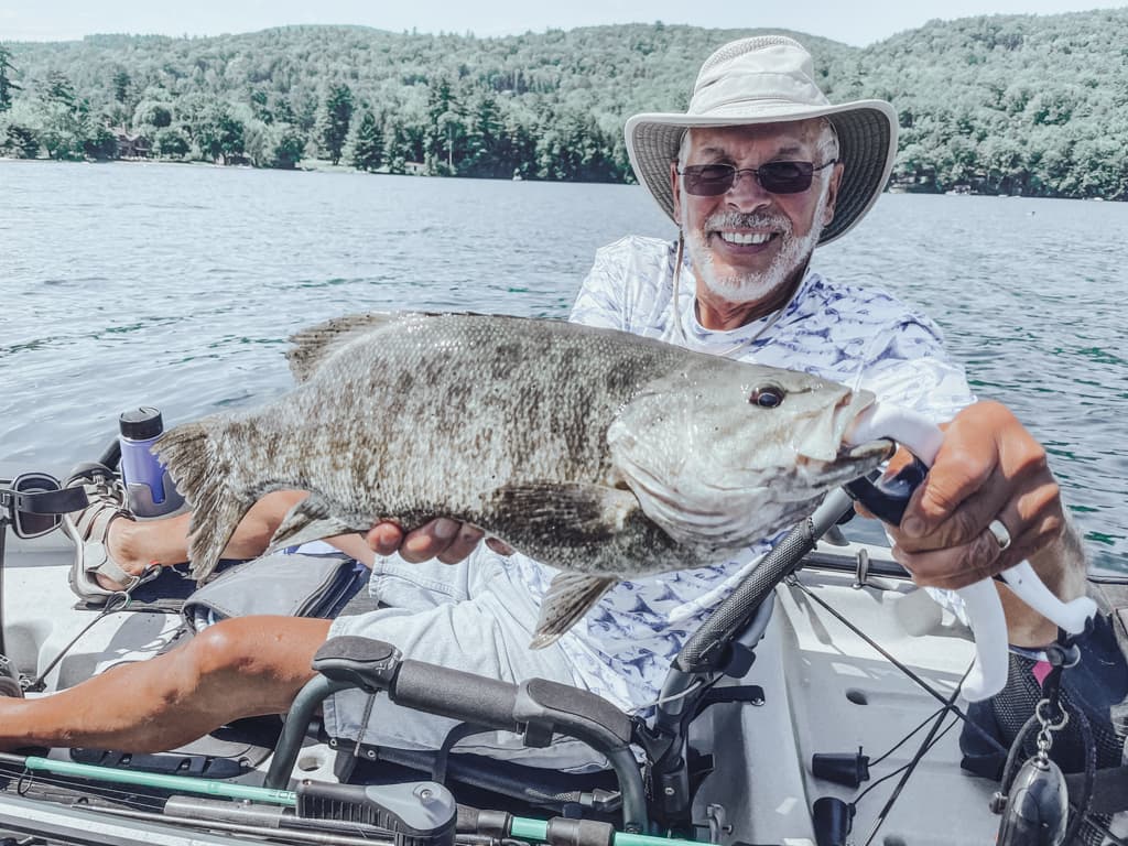 Man in hat and patterned shirt holding one fish while smiling in kayak