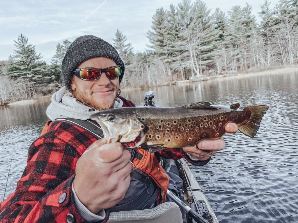 Middle-aged man in a red-and-black flannel and black hat holding a fish on a kayak while smiling