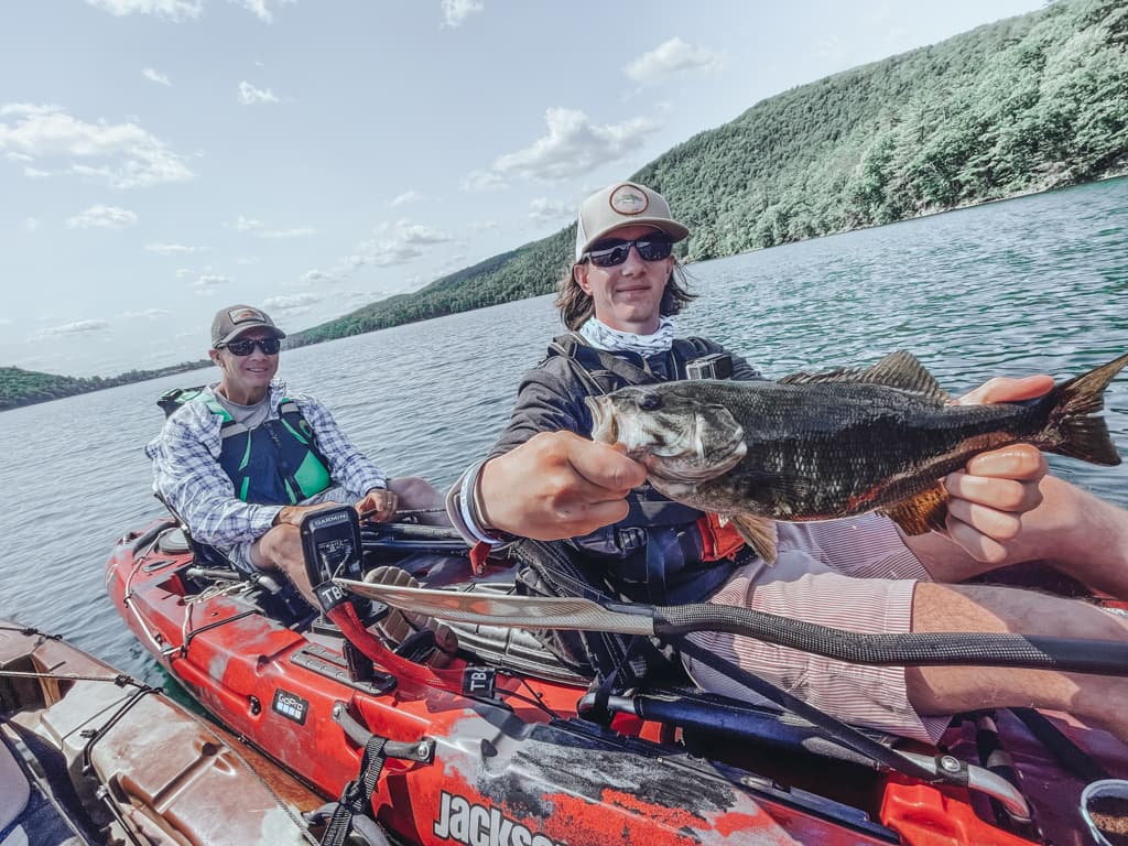 Two men on a kayak; one man on the right is holding a fish and one man on the left is sitting beside him