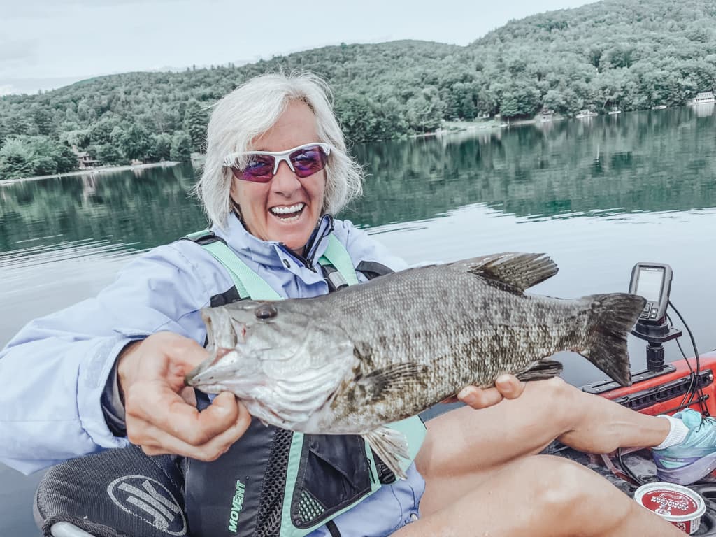 Woman in purple jacket holding one fish while smiling in kayak