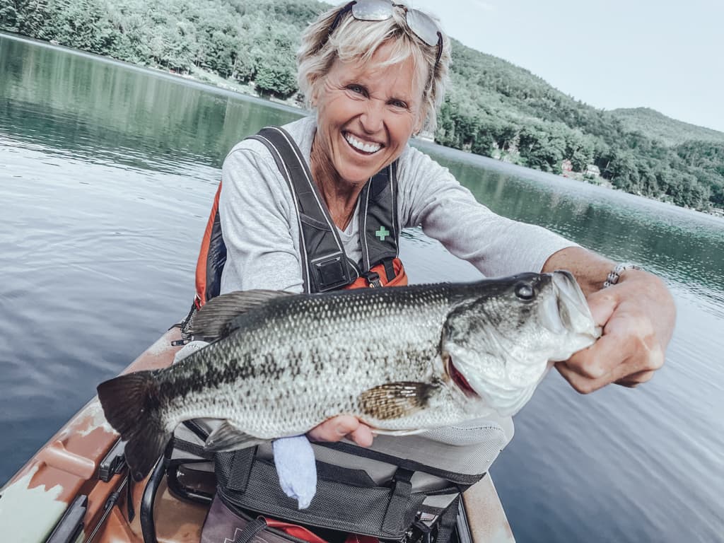 One woman on the left in a kayak while smiling and holding a fishing rod; one woman on the right in a kayakk while smiling and holding a fish