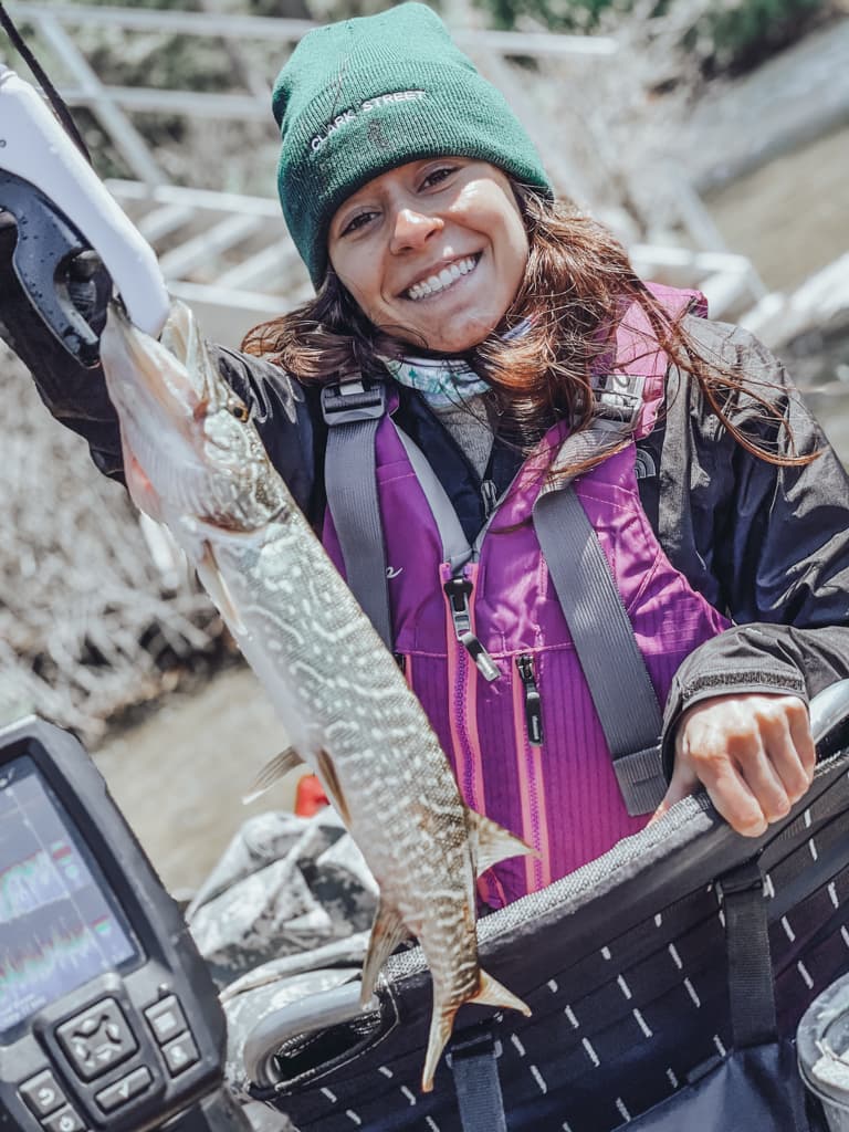 Young woman in a black jacket and purple life-kacjet holding one fish in a kayak while smirking