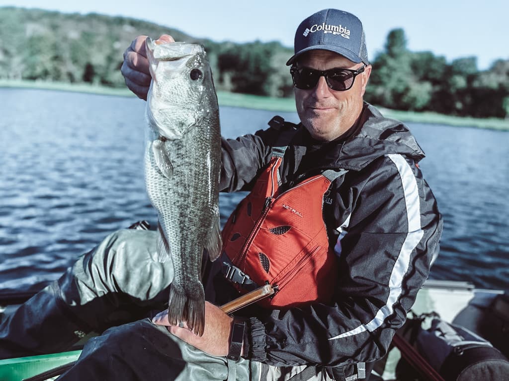 Man in a black jacket and navy hat holding one fish while smiling in a kayak