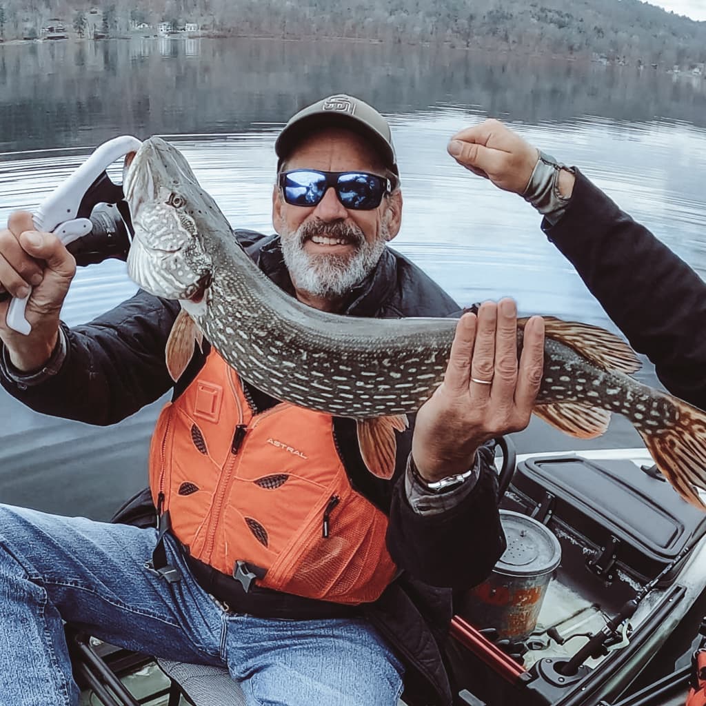 Man in a black jacket, San Diego hat, and light blue jeans holding one fish while smiling in a kayak