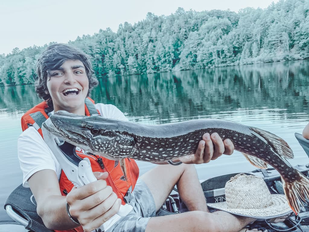 Young man wearing a white shirt and a red life-jacket holding a fish while smiling on a kayak