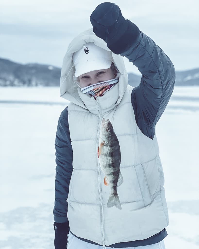 Woman wearing black jacket, white vest, white hat, and black gloves holding a fish on a frozen lake