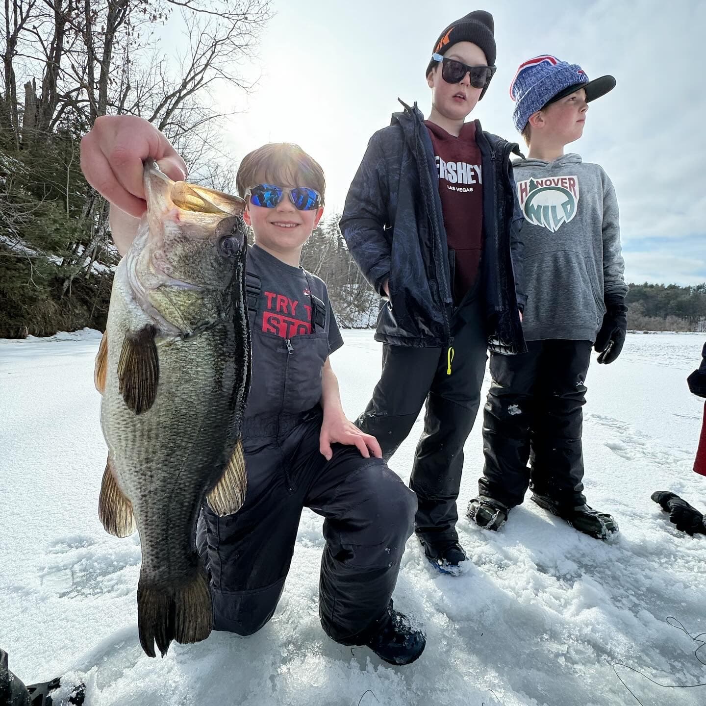 Boy kneeling on frozen lake while holding fish vertically with two other boys to his right