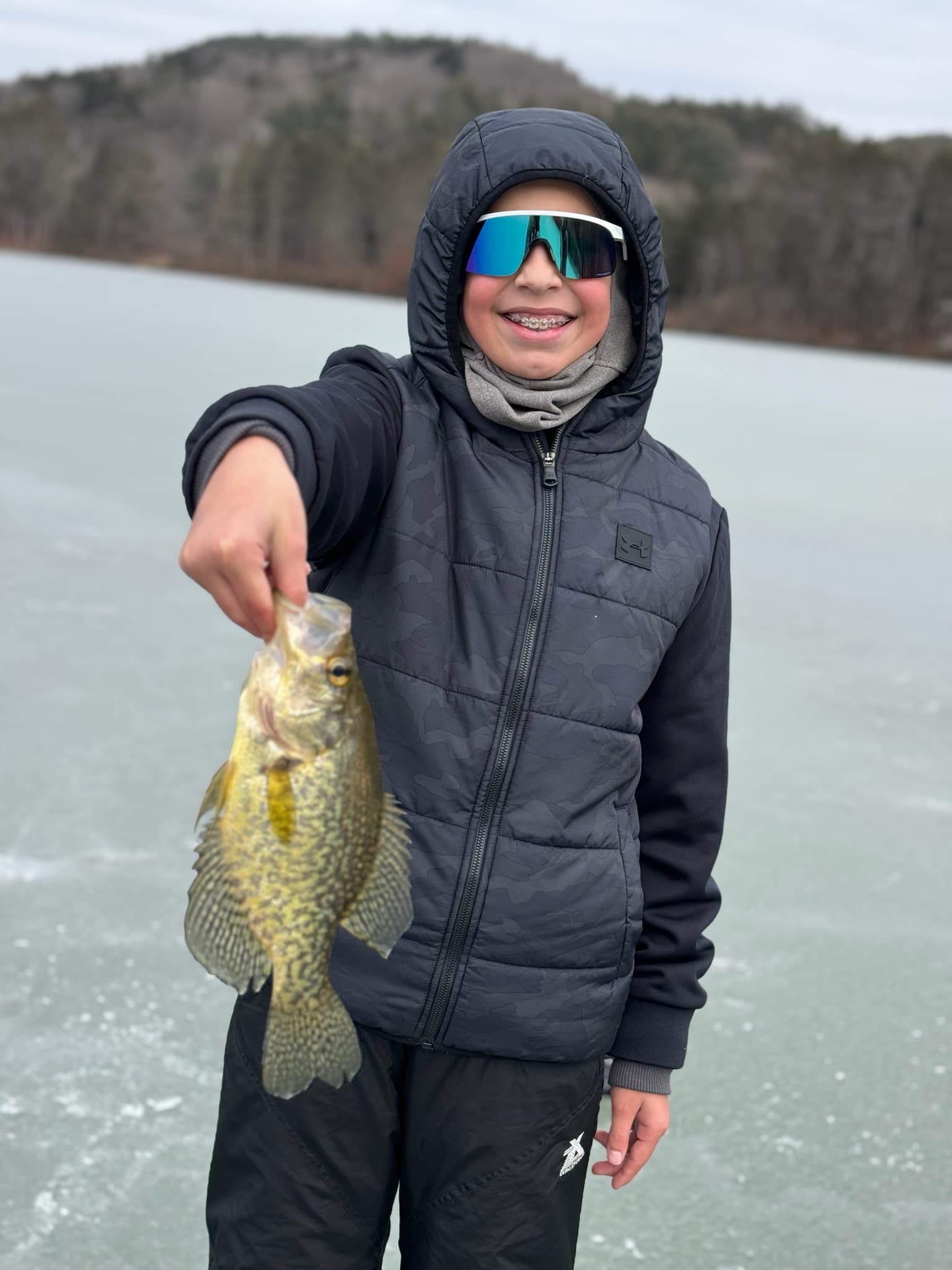 Boy in visor sunglasses holding fish vertically while standing on frozen lake