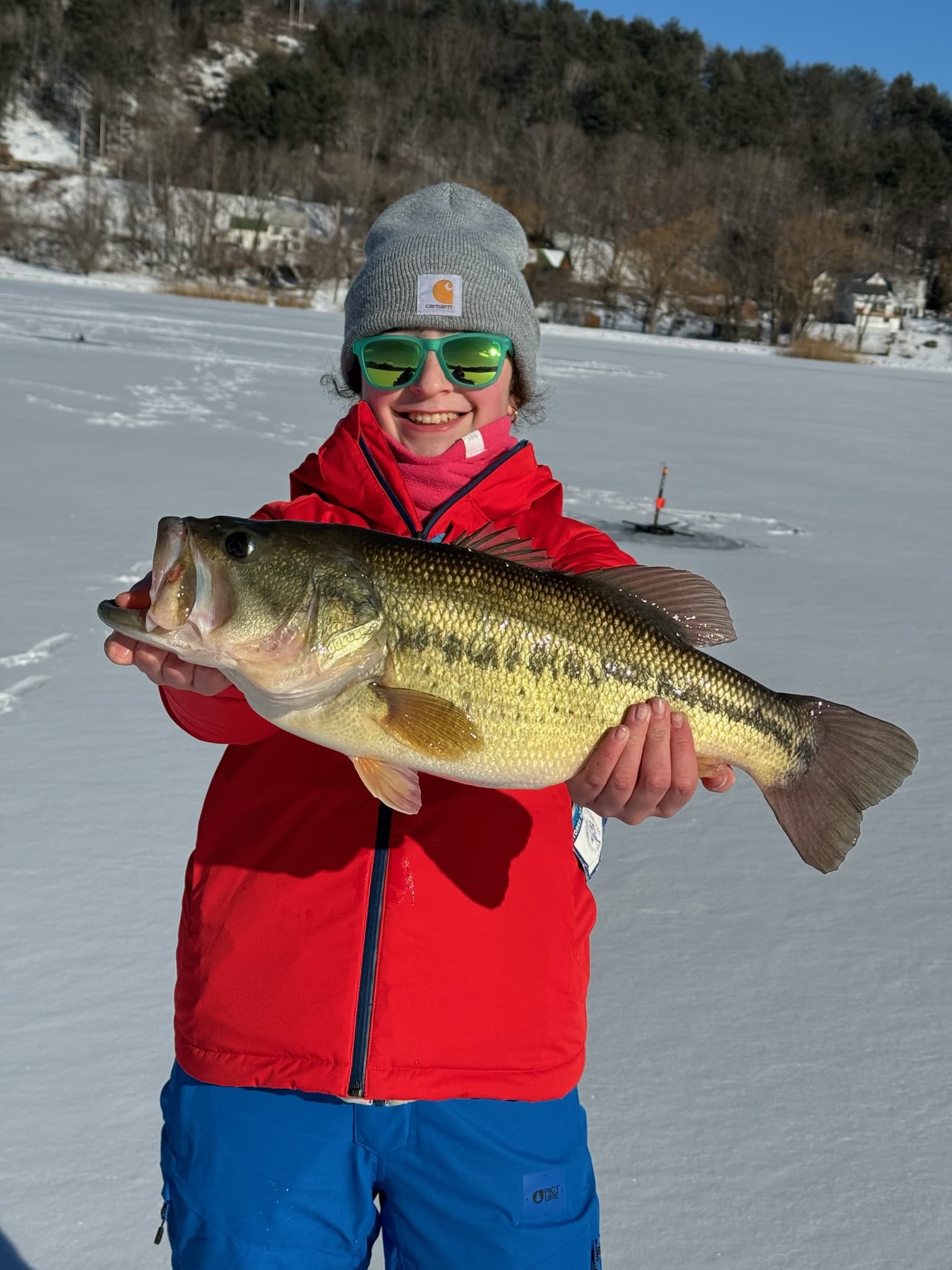 Girl in green sunglasses, red jacket, and grey hat holding fish horizontally while standing on frozen lake