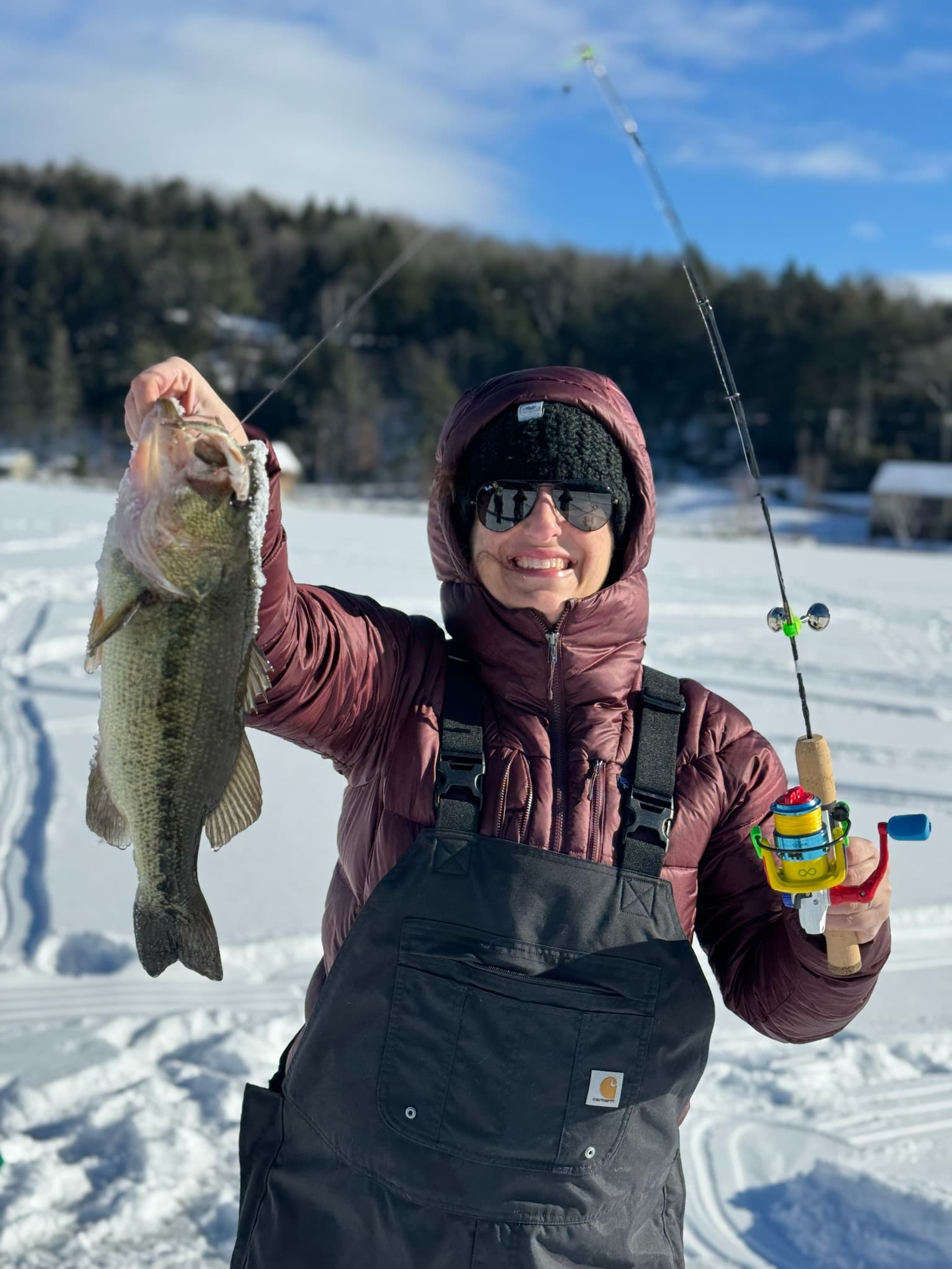 Woman in maroon puffer jacket and black bib holding fishing rod and fish while standing on lake