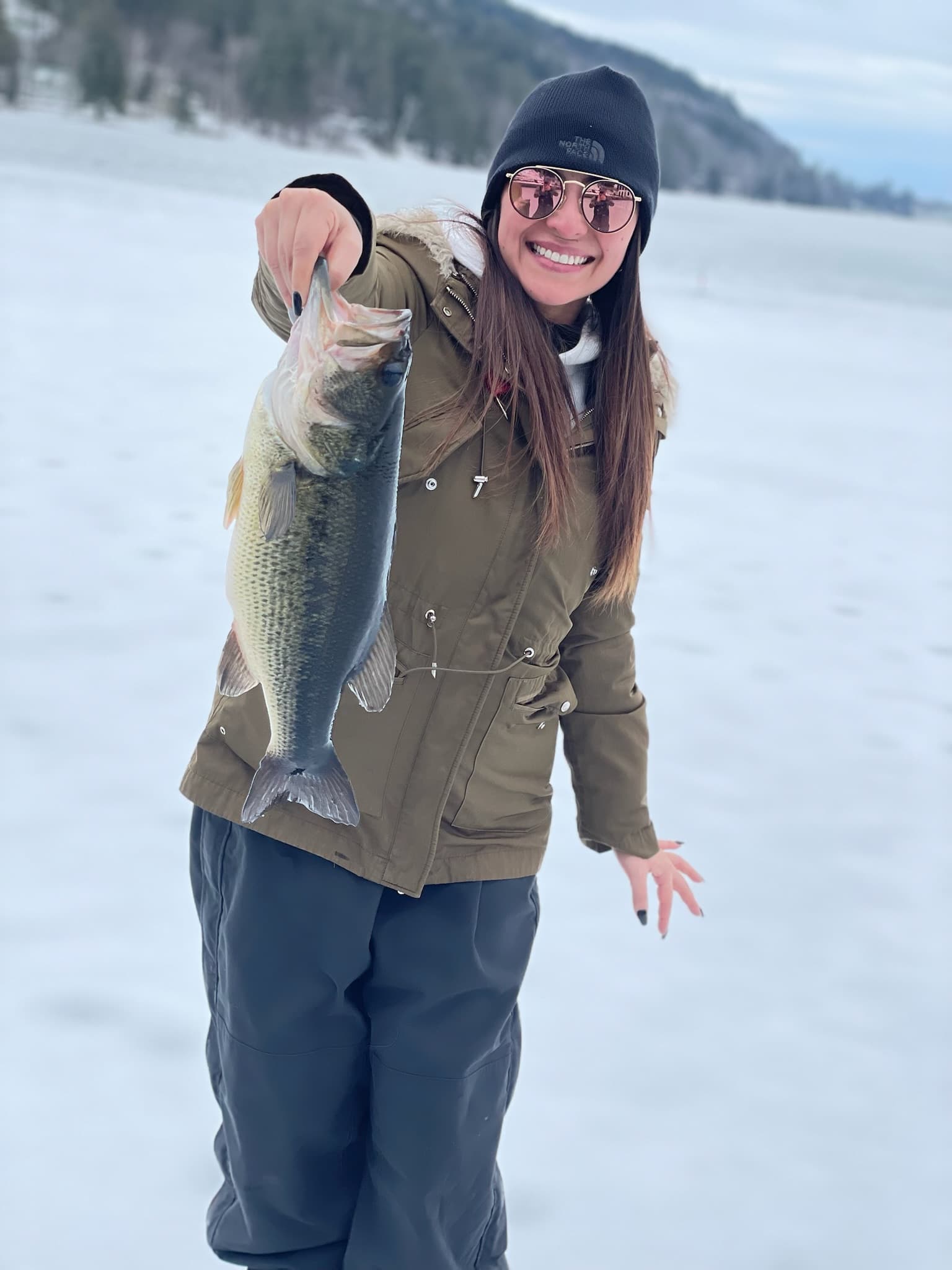 Woman in green jacket and black north face hat smiling while holding fish vertically on frozen lake