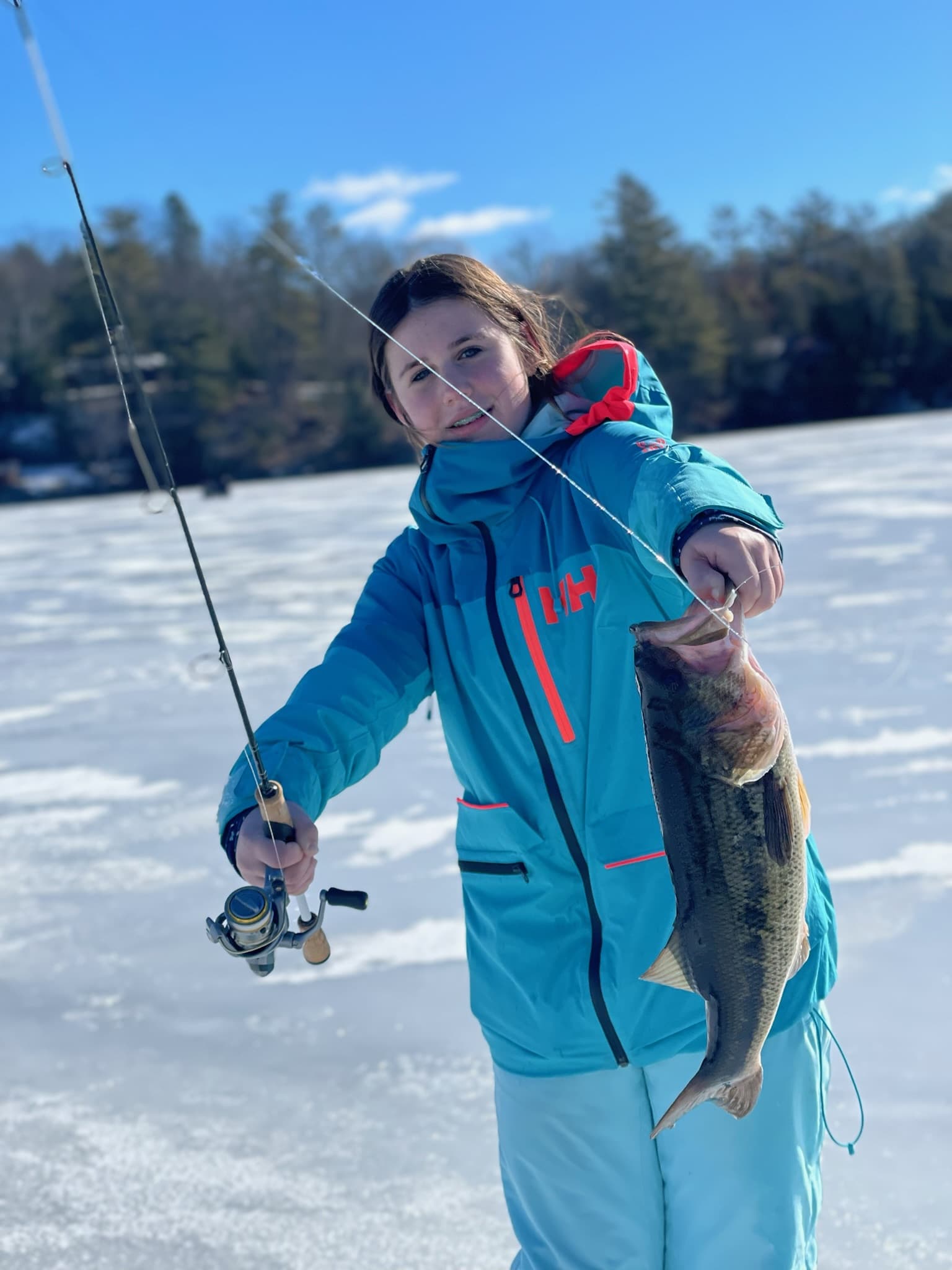 Teenage girl in blue ski jacket and light blue snow pants holding fishing rod and fish while standing on lake