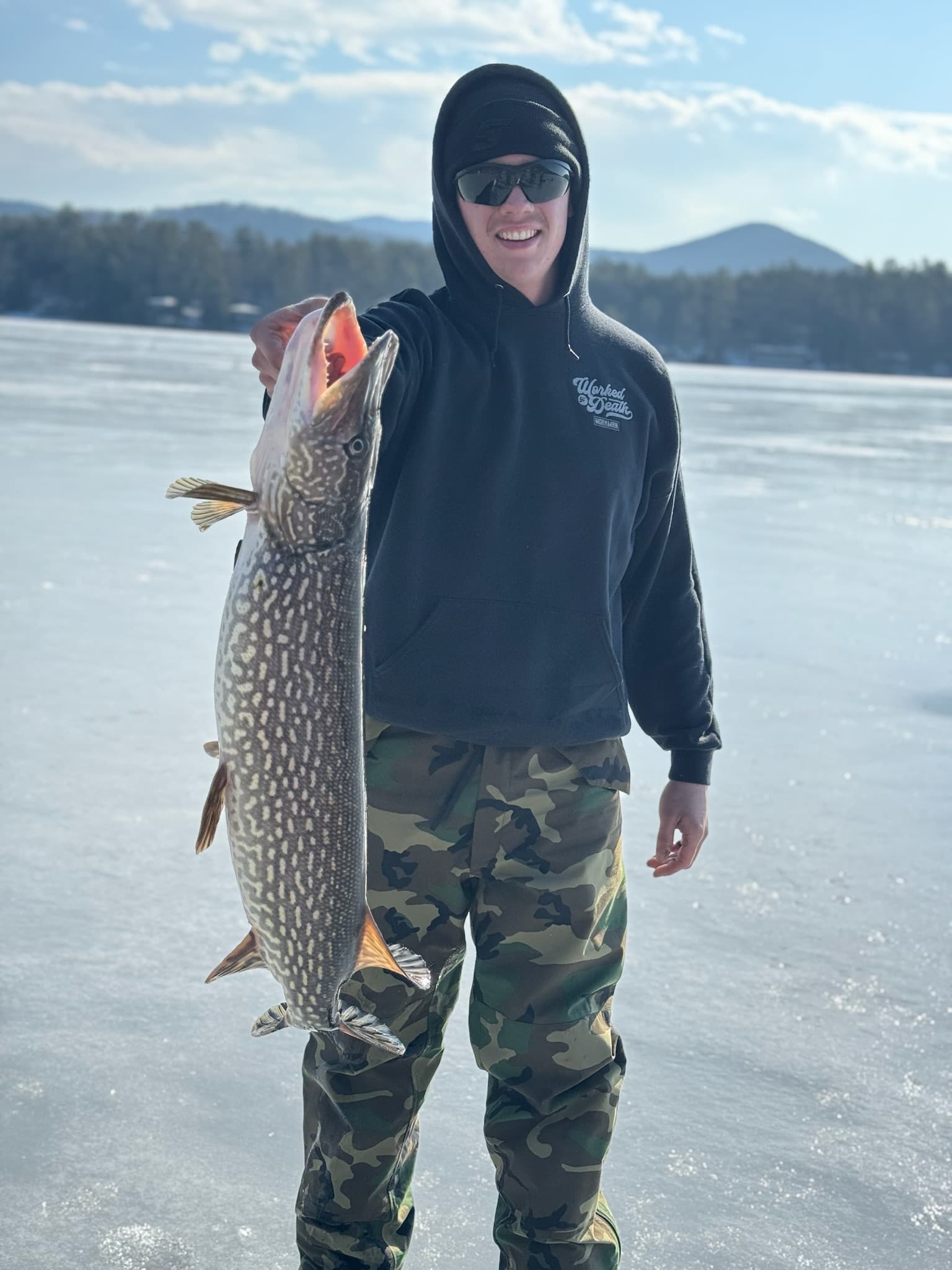 Man in camo pants, black sweatshirt, and black hat holding a fish verticallly while standing on lake