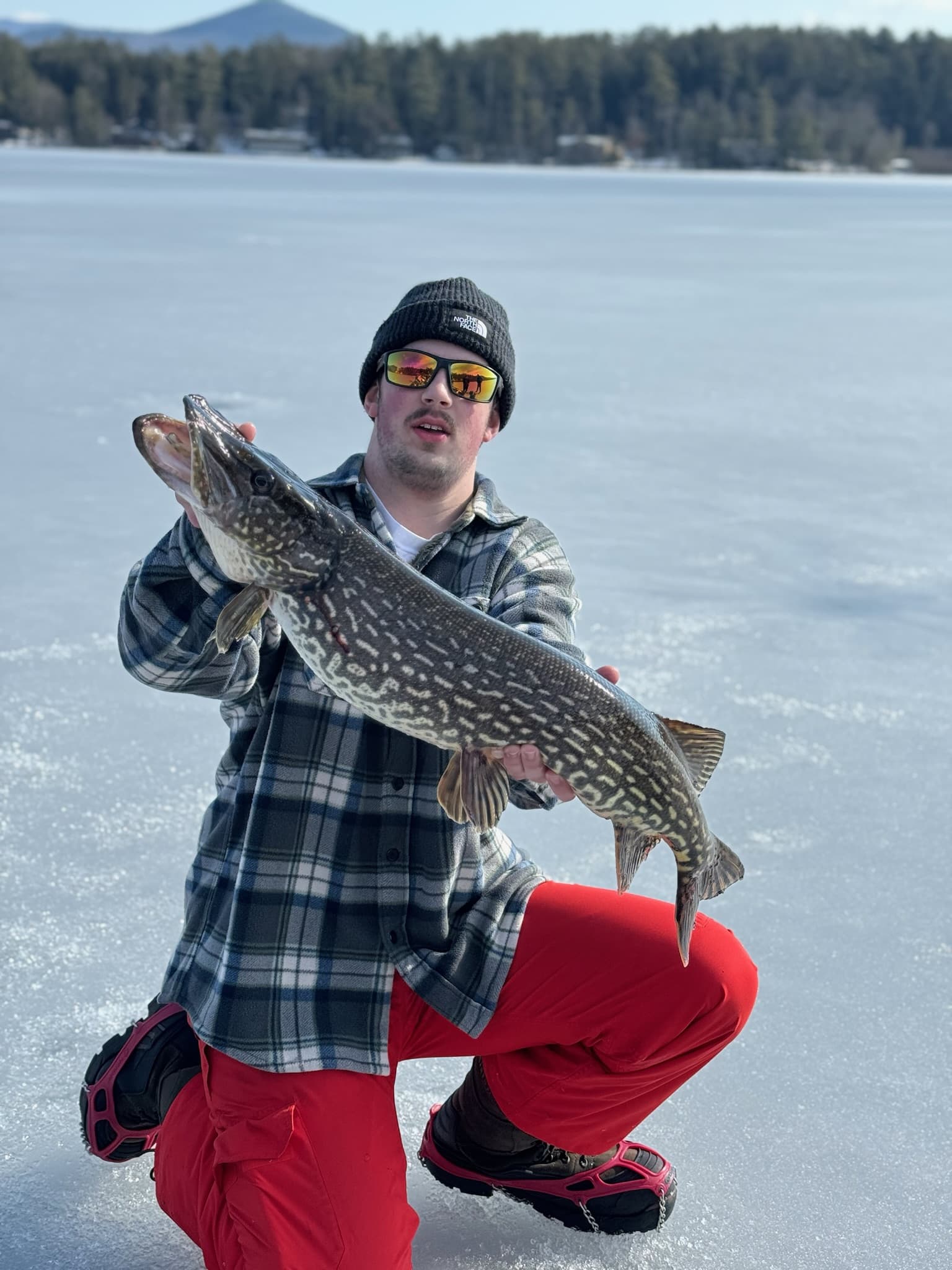 Man in red snow pants, grey flannel, and black hat holding fish horizontally while kneeling on lake
