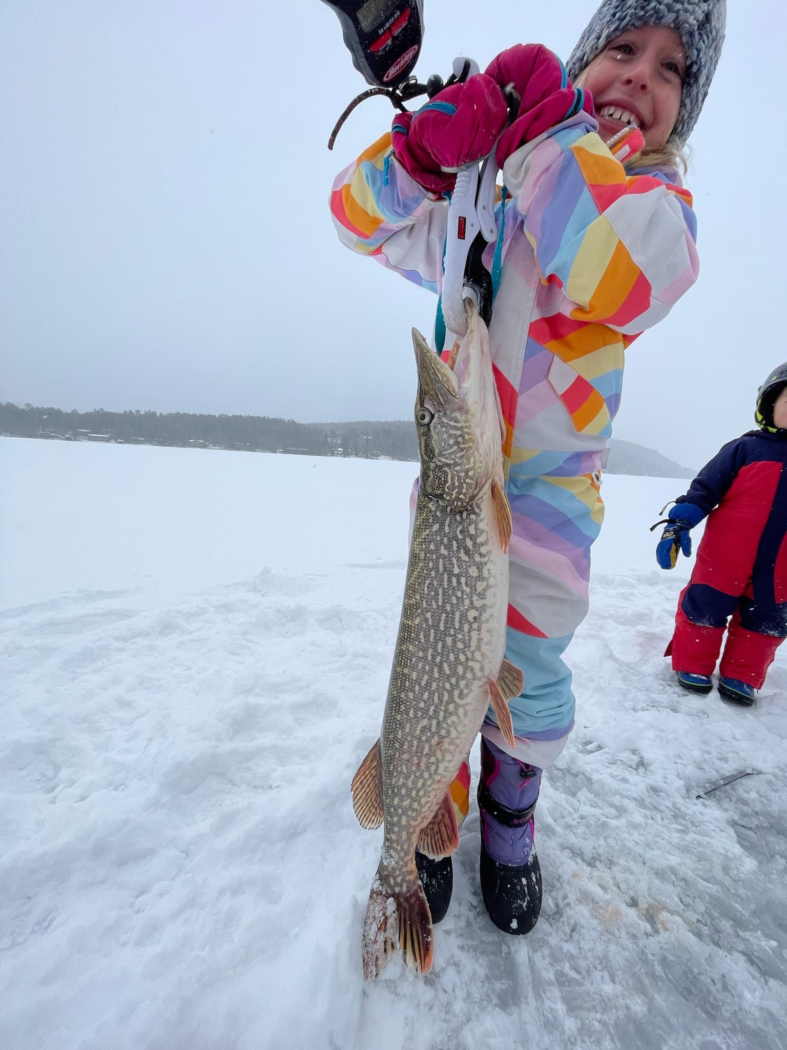 Girl in snowsuit holding fish vertically while standing on frozen lake