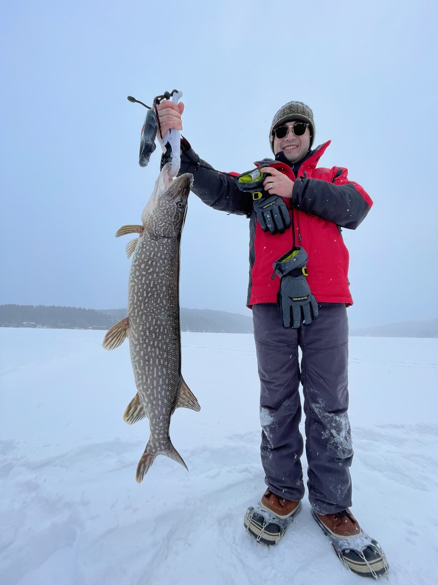 Boy in red jacket and grey snow pants smiling while holding fish vertically on frozen lake