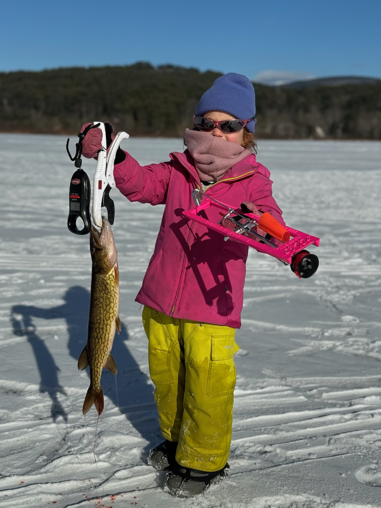 Girl in pink jacket, blue hat, and yellow snow pants holding fish vertically while standing on frozen lake