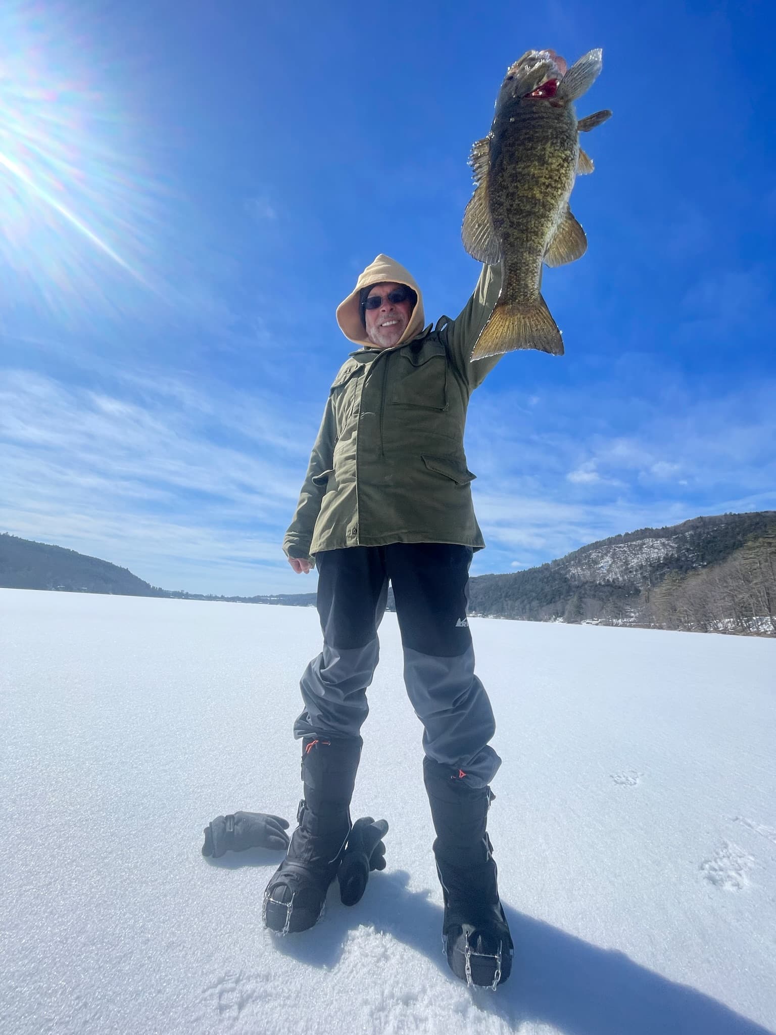 Older man in green jacket and black pants smiling while holding fish vertically on frozen lake