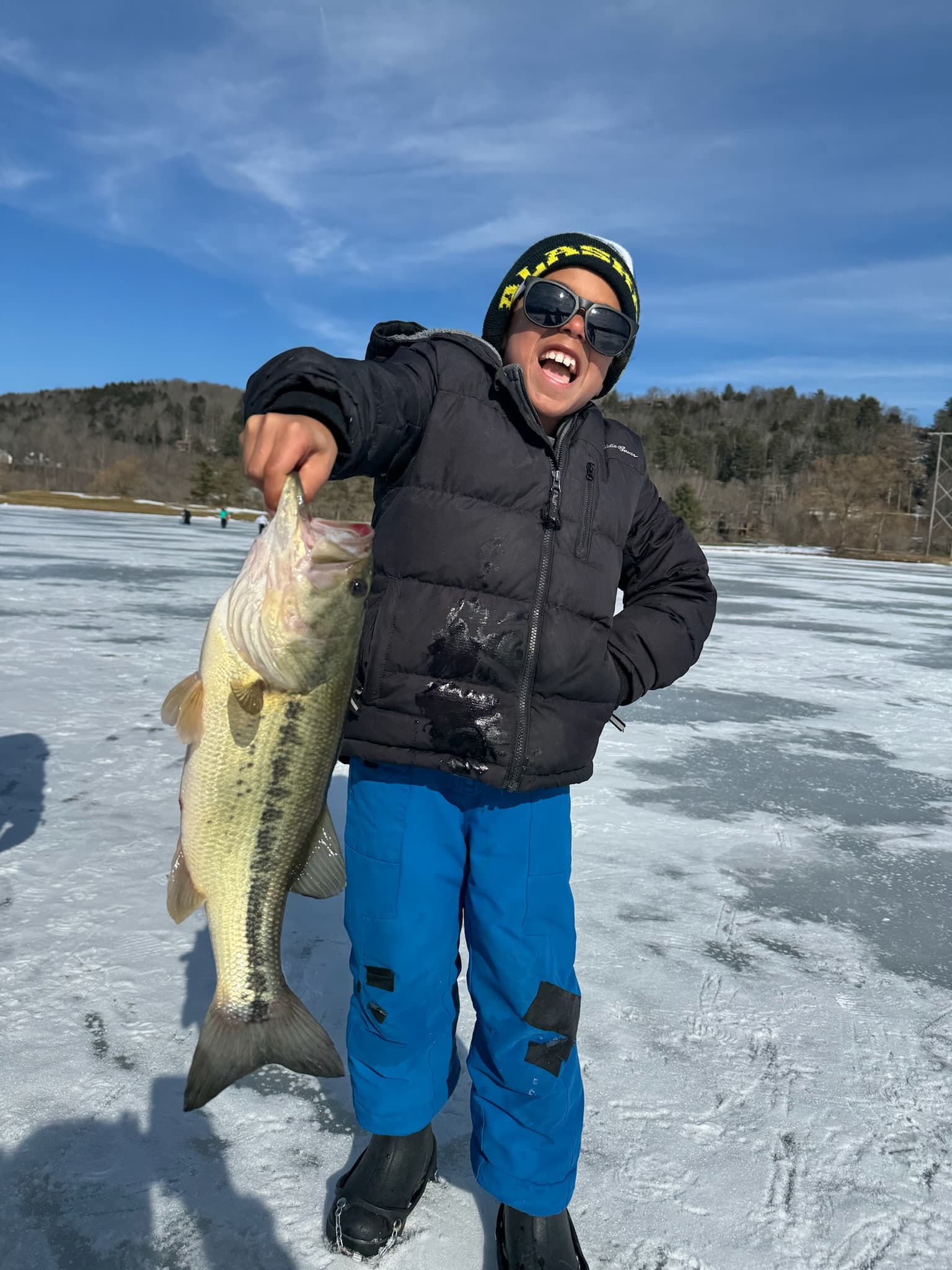 Boy in black puffer jacket and blue snow pants holding fish vertically on frozen lake