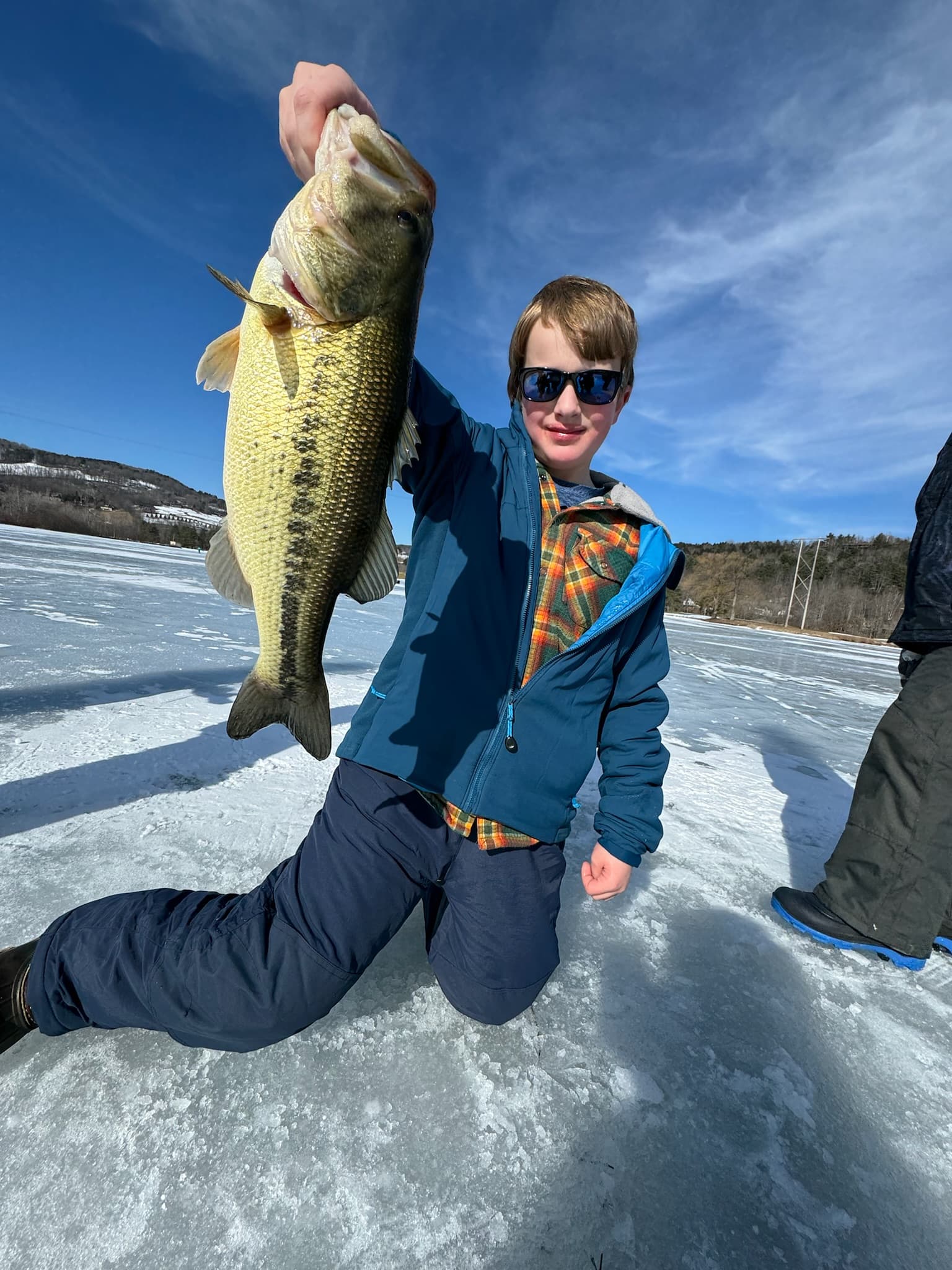 Boy in blue jacket and navy snow pants kneeling on frozen lake while holding fish vertically