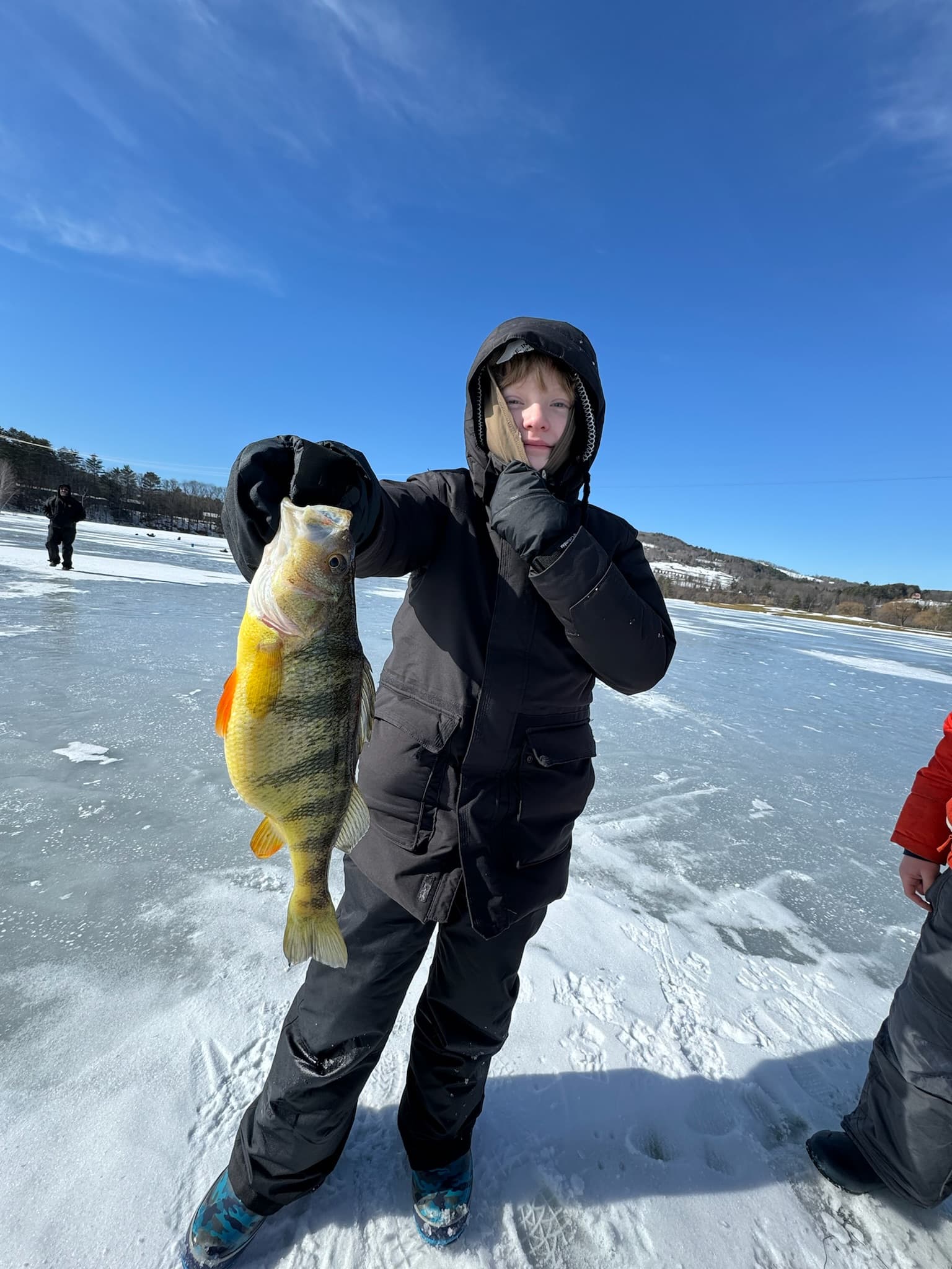 Boy in black jacket and snow pants holding fish vertically on frozen lake