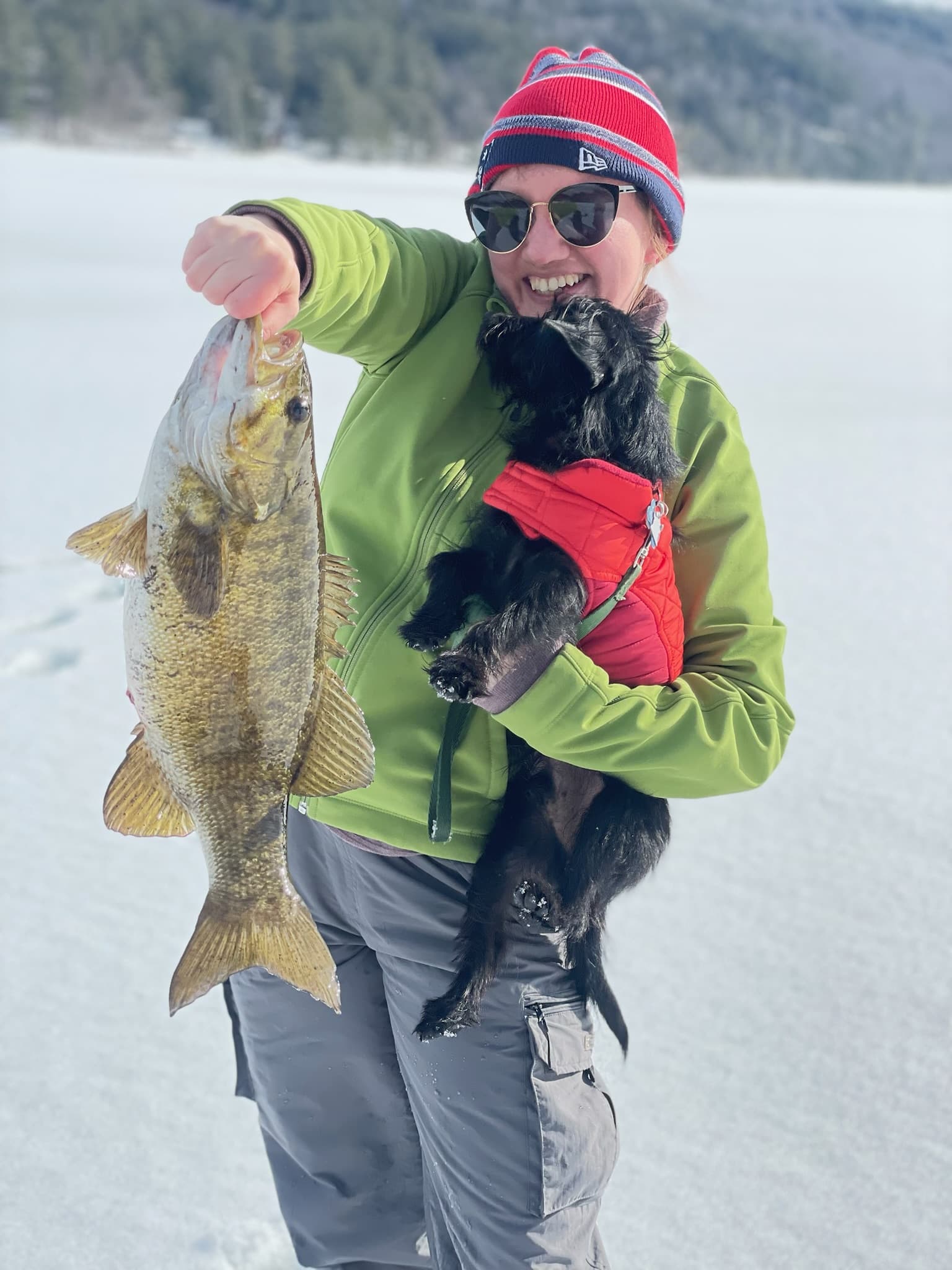 Woman holding small black dog in arms and fish vertically in other hand on frozen lake