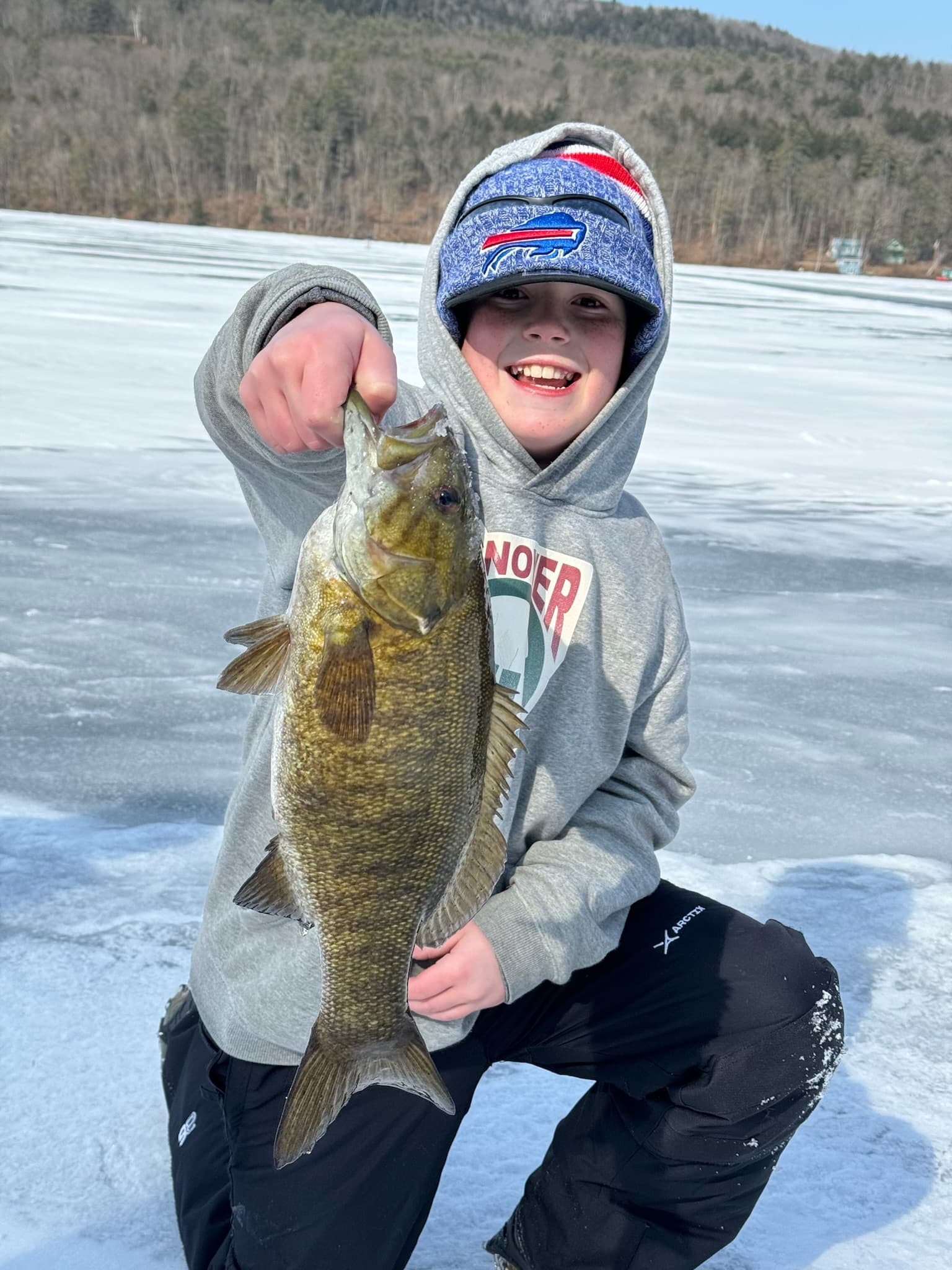 Boy in grey sweatshirt and black snow pants kneeling while holding fish vertically on frozen lake