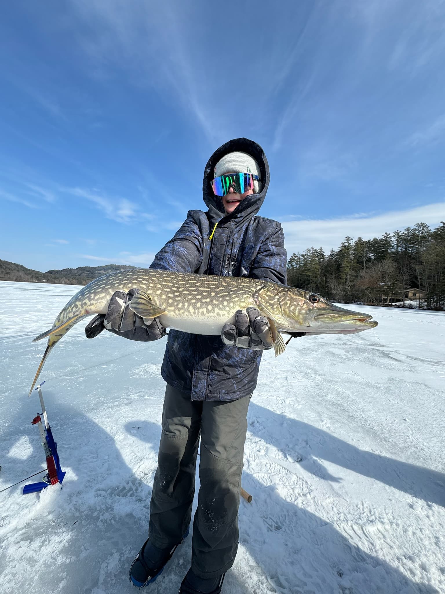 Boy in visor sunglasses, navy ski jacket, and grey snow pants holding fish horizontally on frozen lake