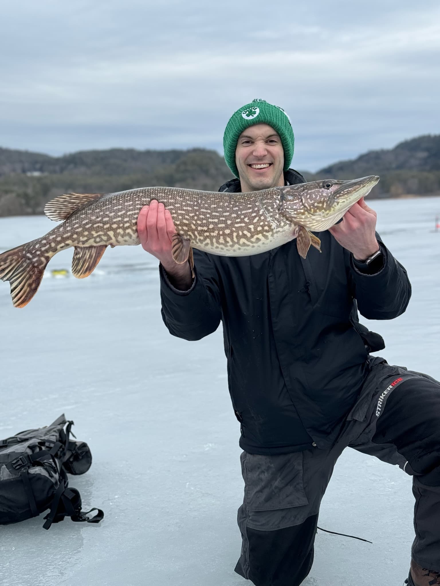 Man in green hat, black snow pants, and black ski jacket smiling and kneeling while holding fish horiztonally on frozen lake