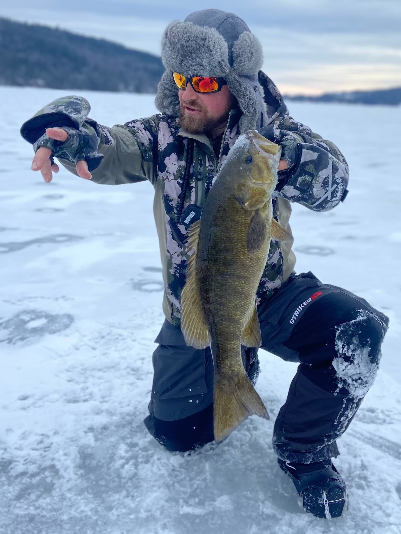 Man in fur hat kneeling on frozen lake while pointing and holding a fish vertically