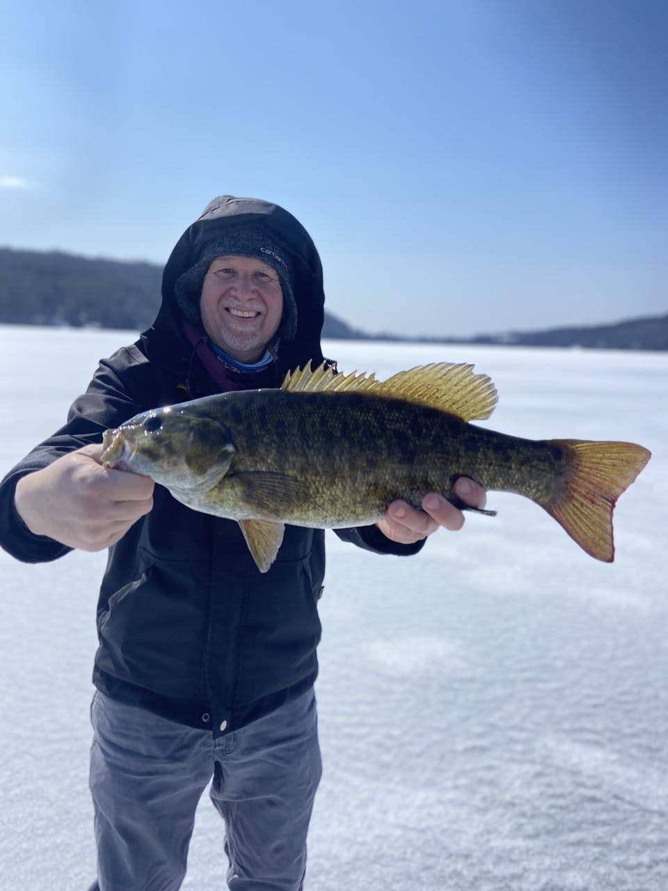 middle-aged man standing on a frozen lake, wearing a winter green jacket with hood up, smiling, and holding a fish horizontally