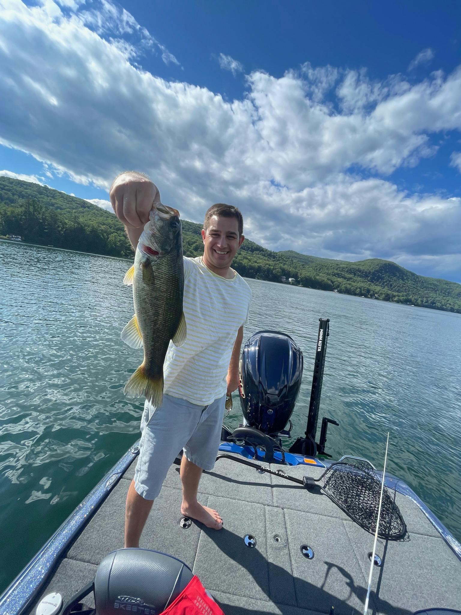 Man with no shoes, grey shorts, and white shirt holds largemouth bass on blue boat