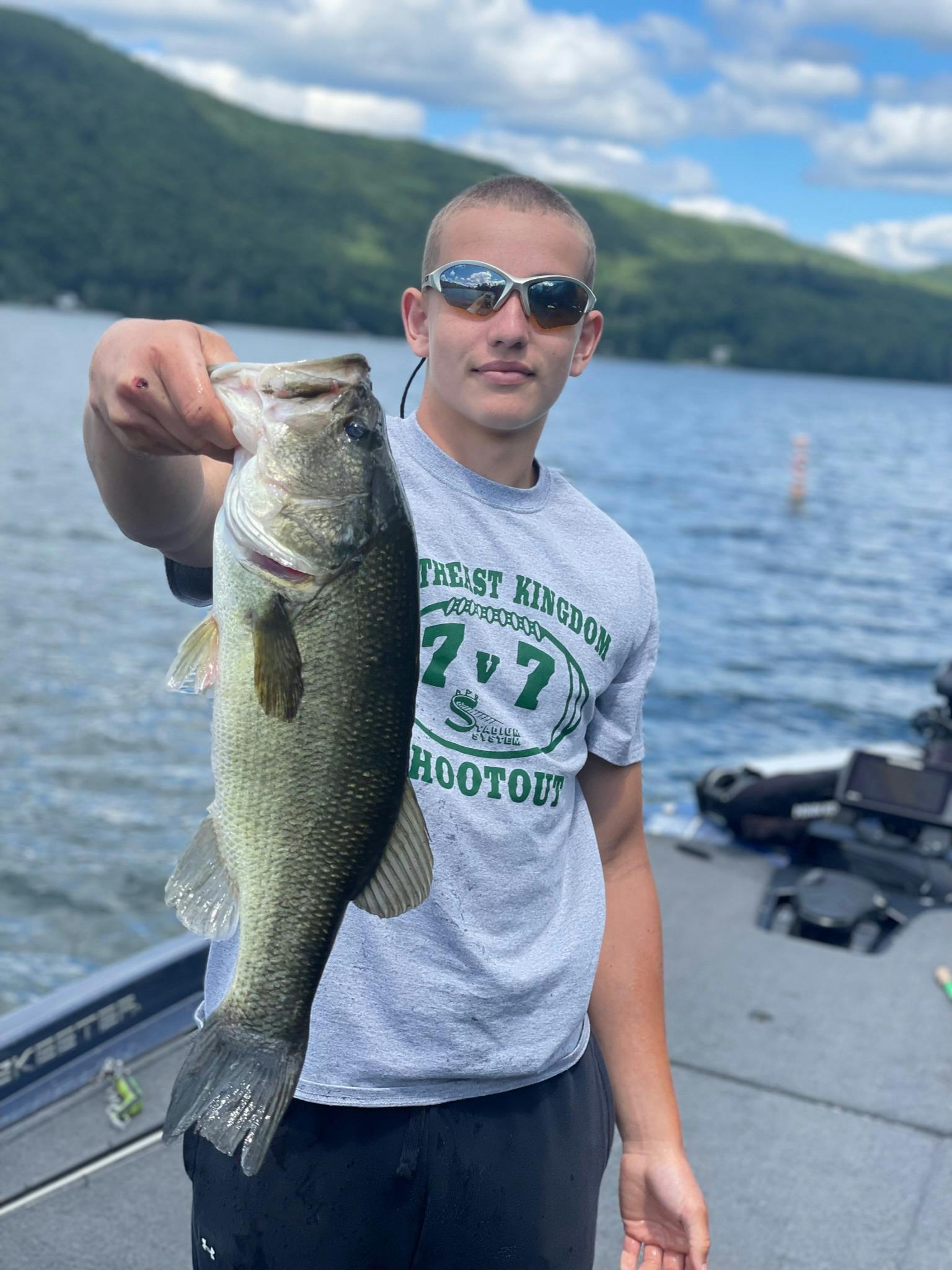 younger man holds largemouth bass on boat while wearing sunglasses, grey shirt, and black shorts