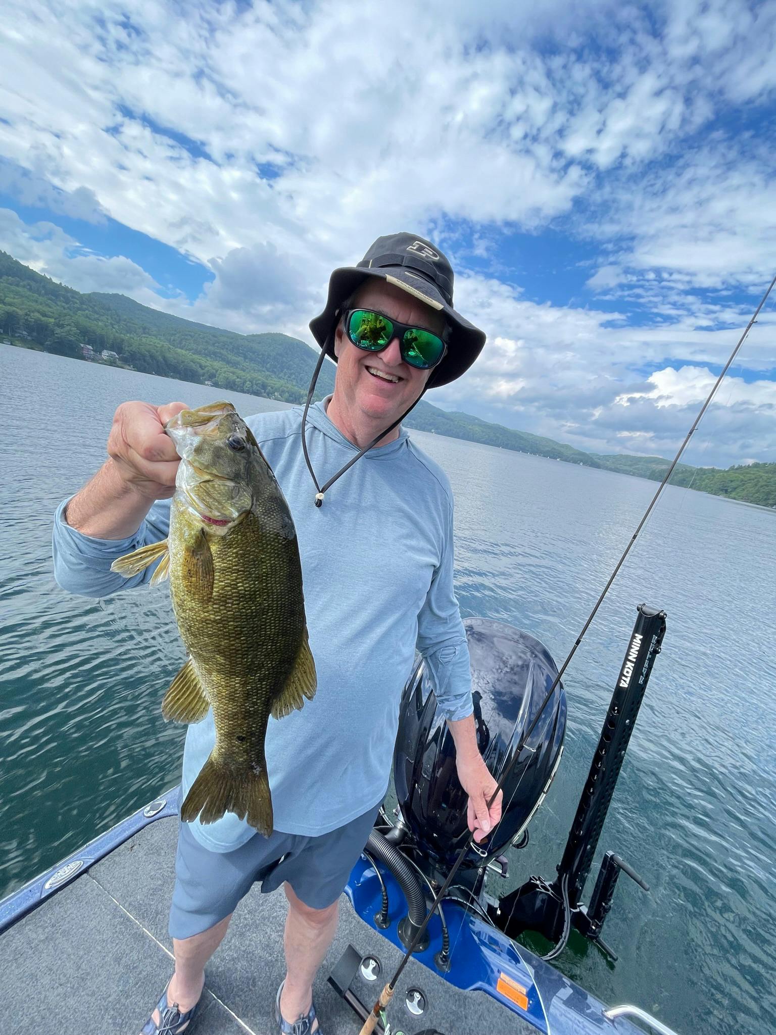 Tall man wearing black bucket hat, blue shirt, and grey shorts holds smallmouth bass on blue boat