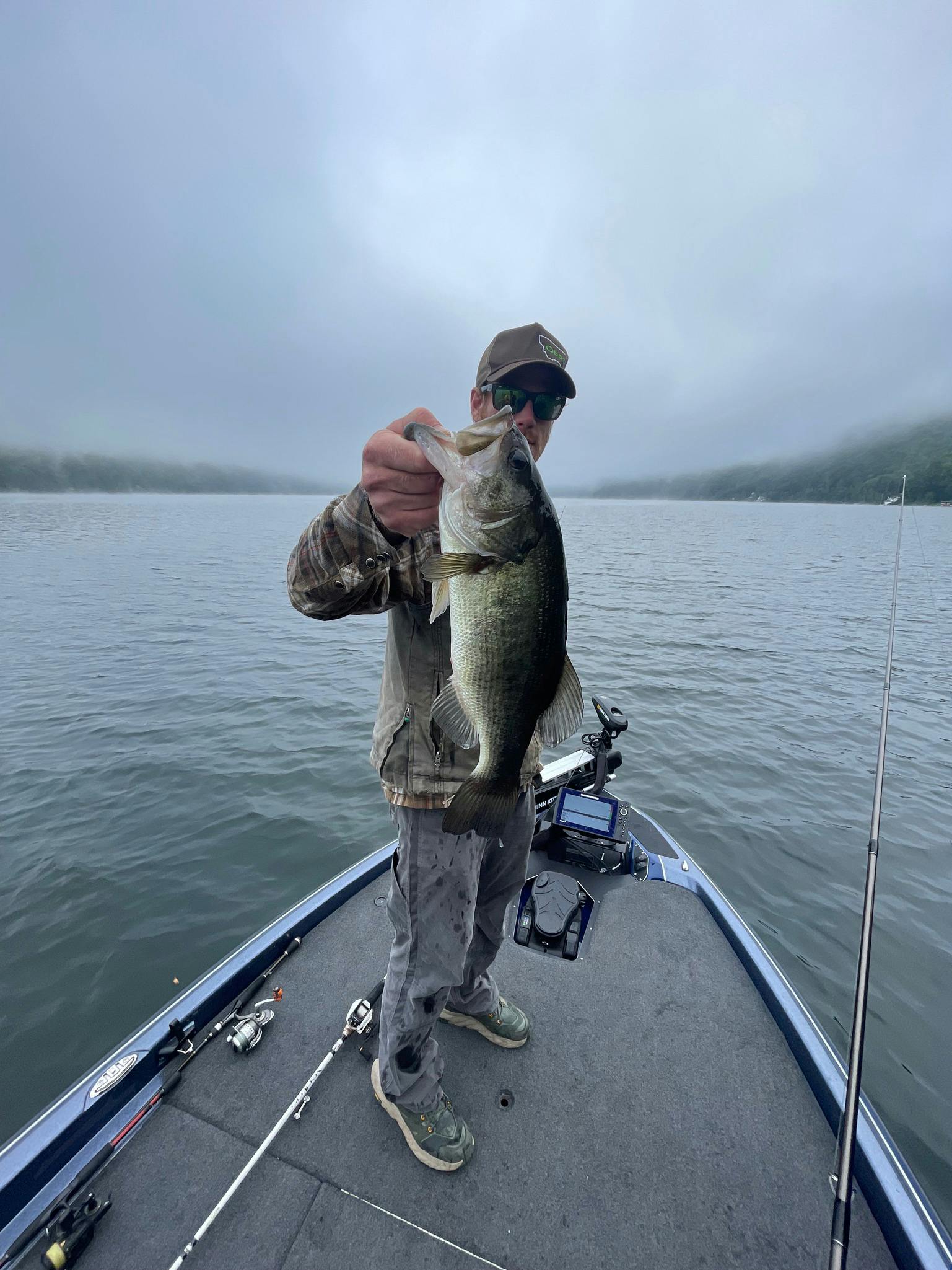 Man in brown baseball cap, camo jacket, and grey pants holds largemouth bass on boat