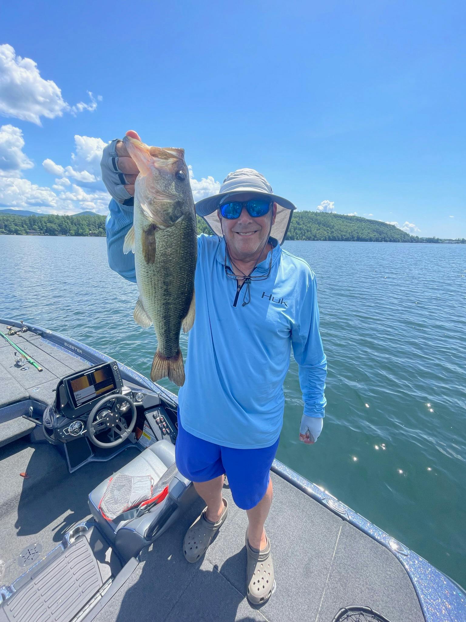 older man in blue shirt, beige crocks, bucket hat, and blue sunglasses holds up largemouth bass on boat