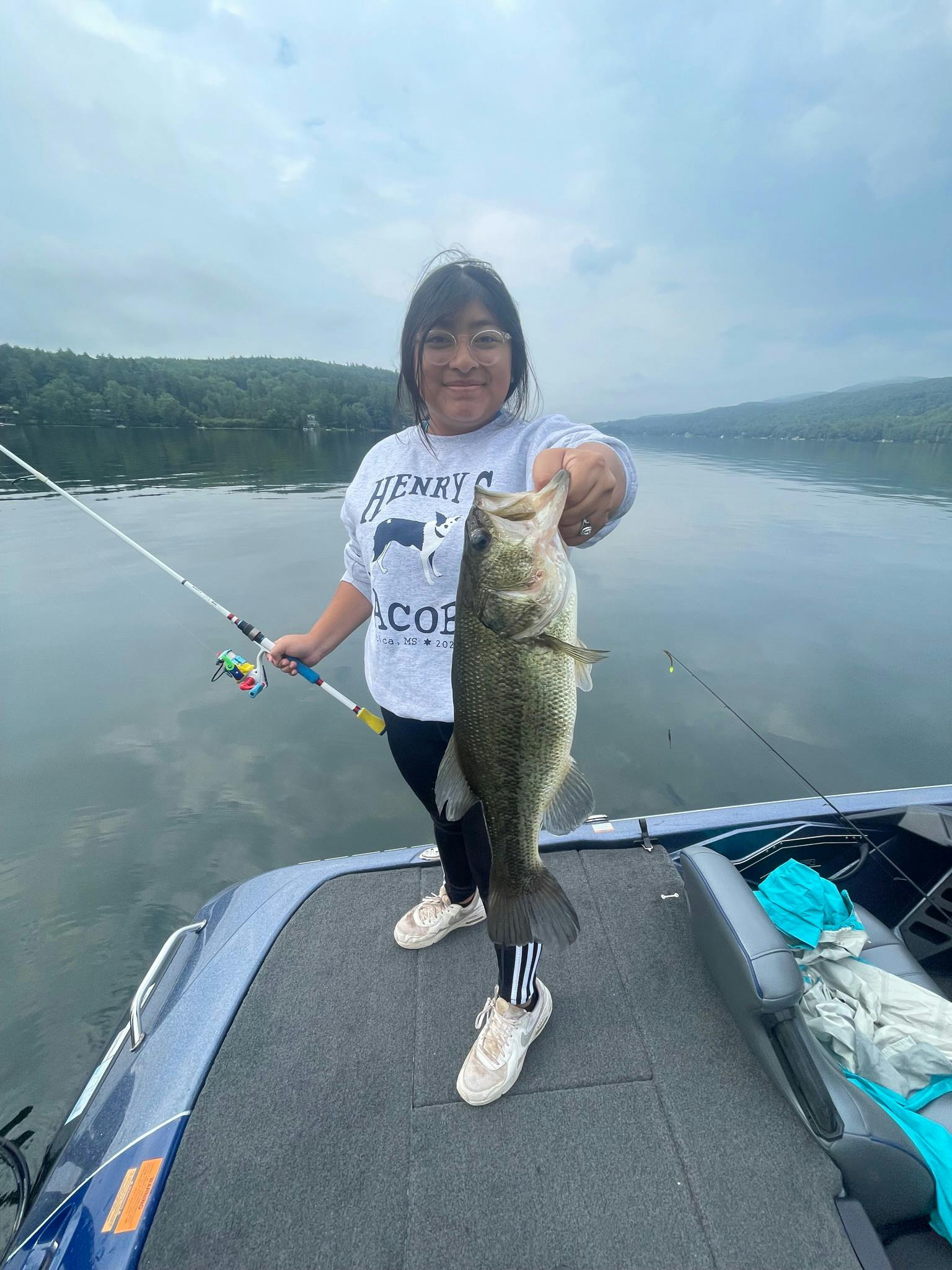 Girl in sweatshirt, black sweatpants, and white shoes holds largemouth bass on blue boat