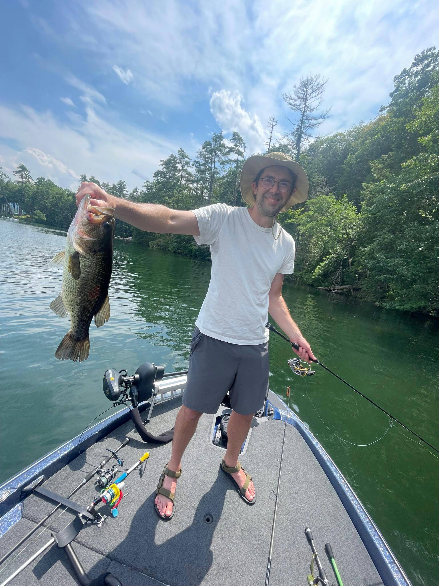 Man in white shirt, grey shorts, and tevas holds largemouth bass on blue boat